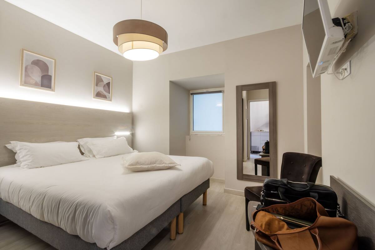 A hotel room with a bed, a desk, a chair, a suitcase, and a mirror.