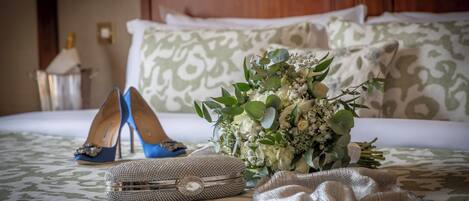 A neatly made bed with patterned bedding, a bouquet of flowers, a pair of blue high heels, and a jeweled clutch on a patterned carpet.