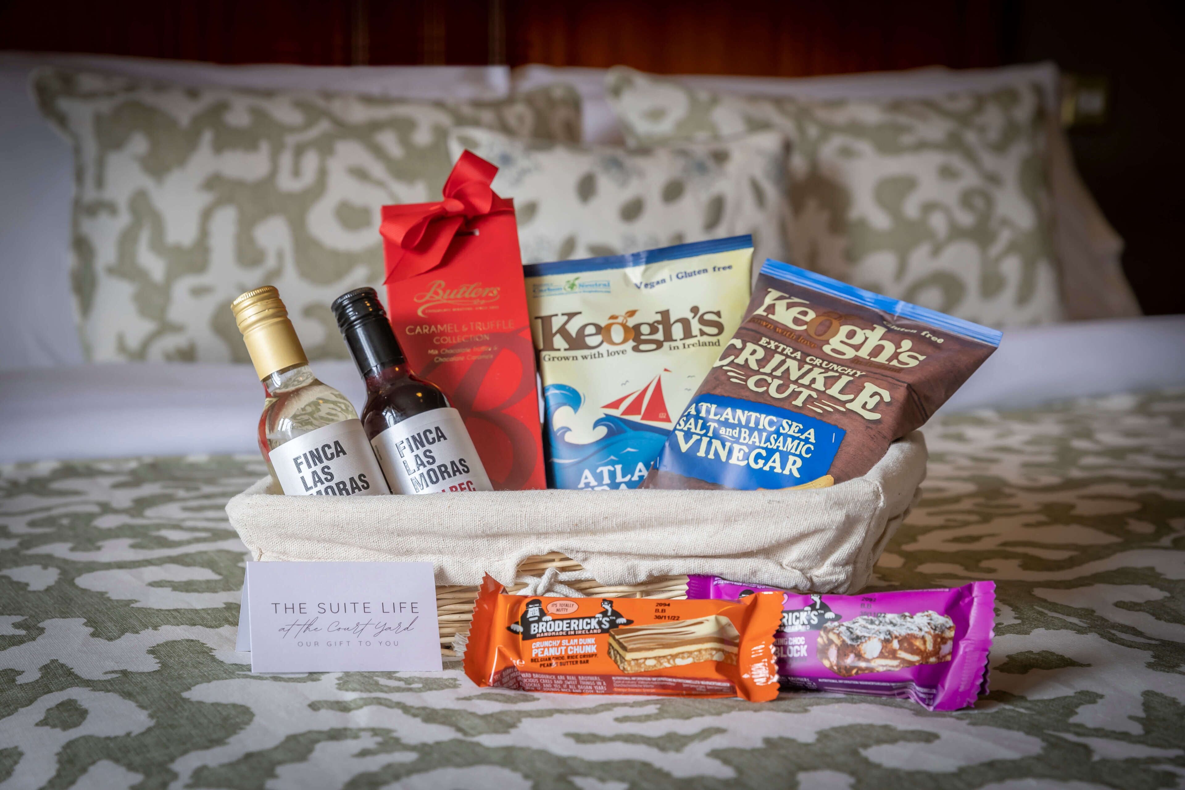 Ein Geschenkset auf einem Bett mit Gegenständen wie einer Weinflasche, Snacks und einem Buch.
