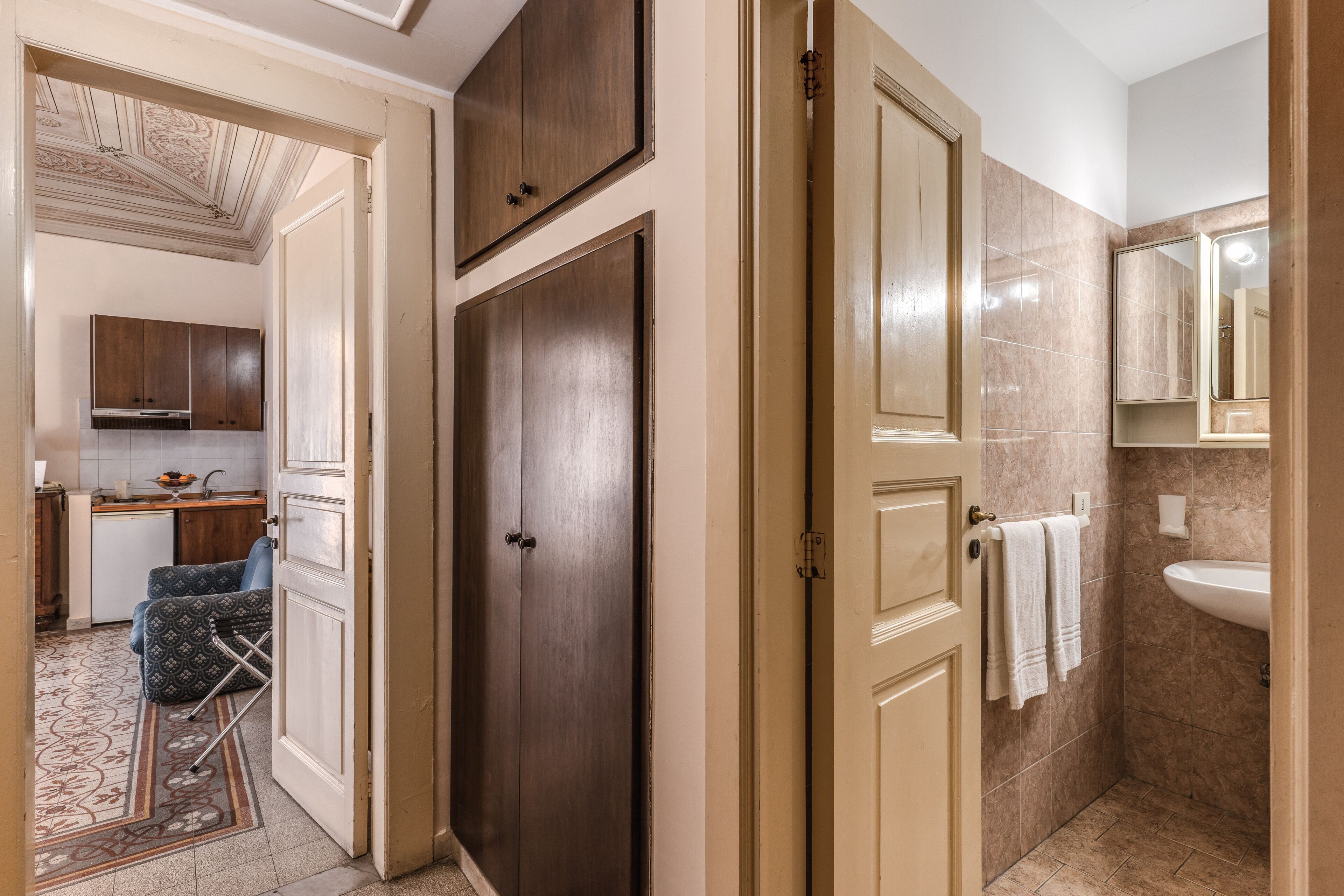 A hotel room with a wooden wardrobe, a bathroom with a sink and mirror, and a kitchen area visible through an open door.