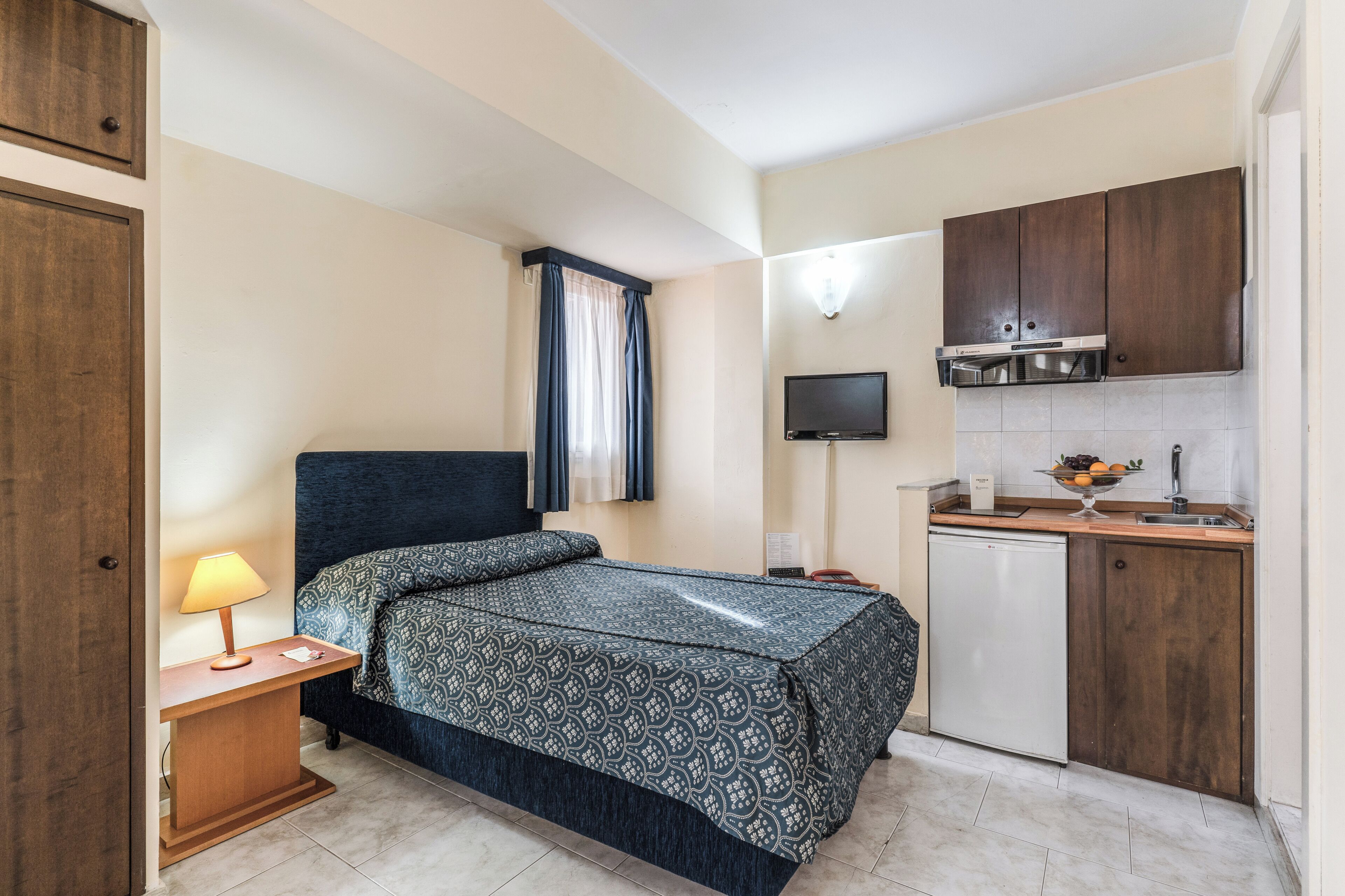 A compact hotel room with a bed, bedside table with a lamp, a small kitchenette with a microwave, and a TV mounted above the counter.