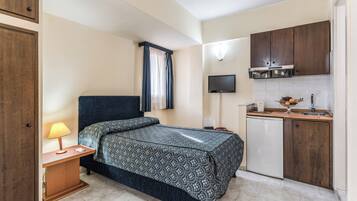 A compact hotel room with a bed, bedside table with a lamp, a small kitchenette with a microwave, and a TV mounted above the counter.
