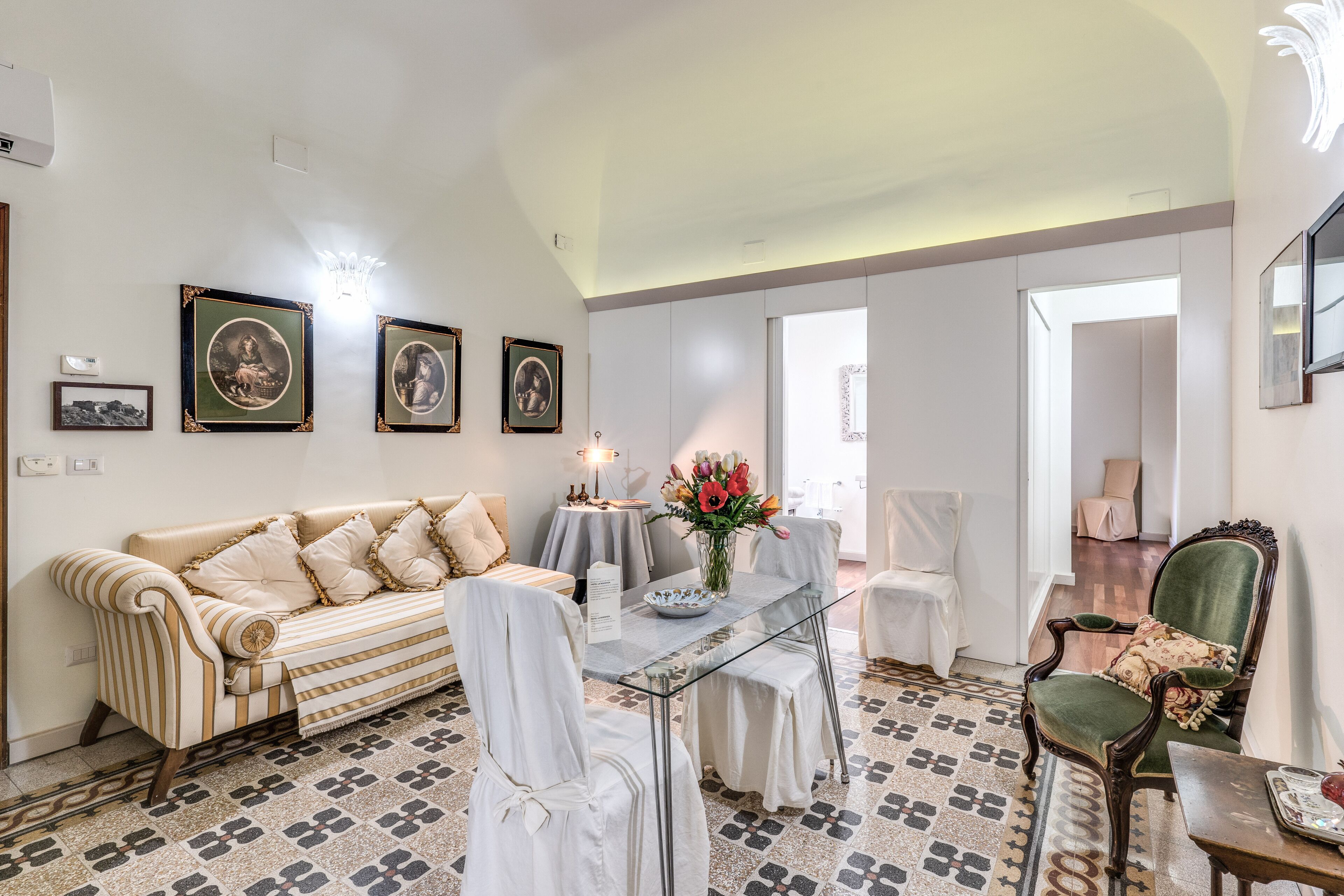 A dining area with a glass table, striped sofa, and chairs with white covers. There are framed pictures on the wall and a vase with flowers on the table.