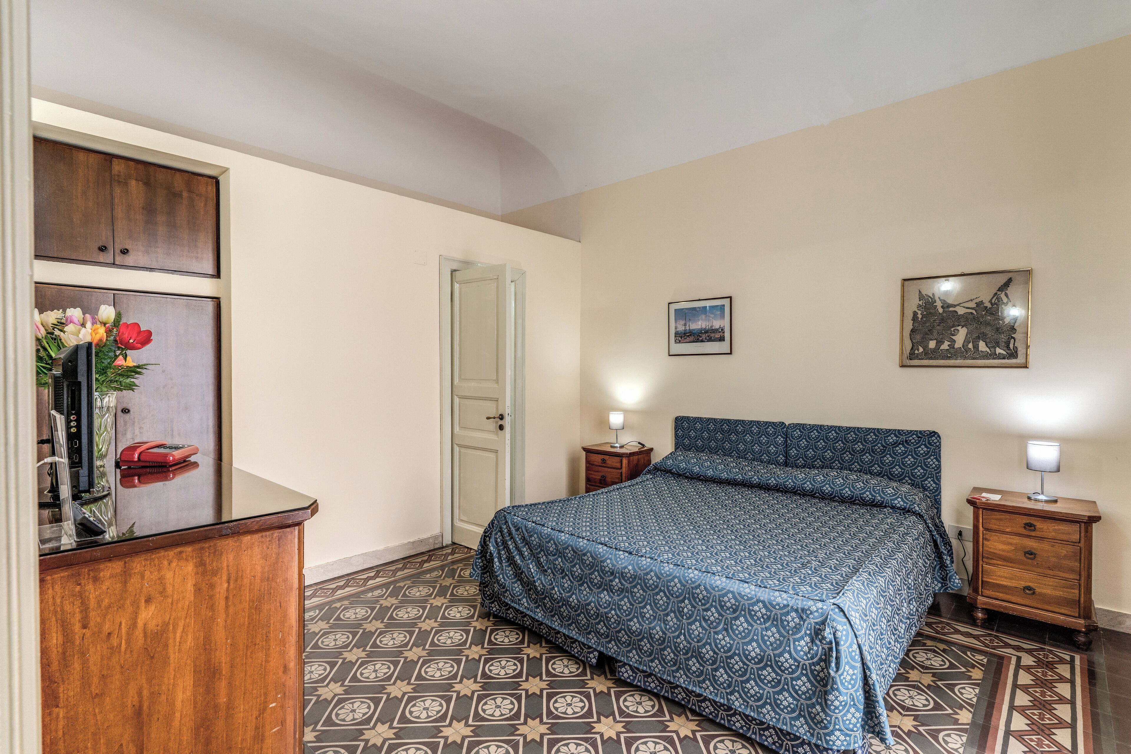 A bedroom with a patterned floor, a bed with a blue patterned bedspread, a wooden dresser, a nightstand with a lamp, and a door.
