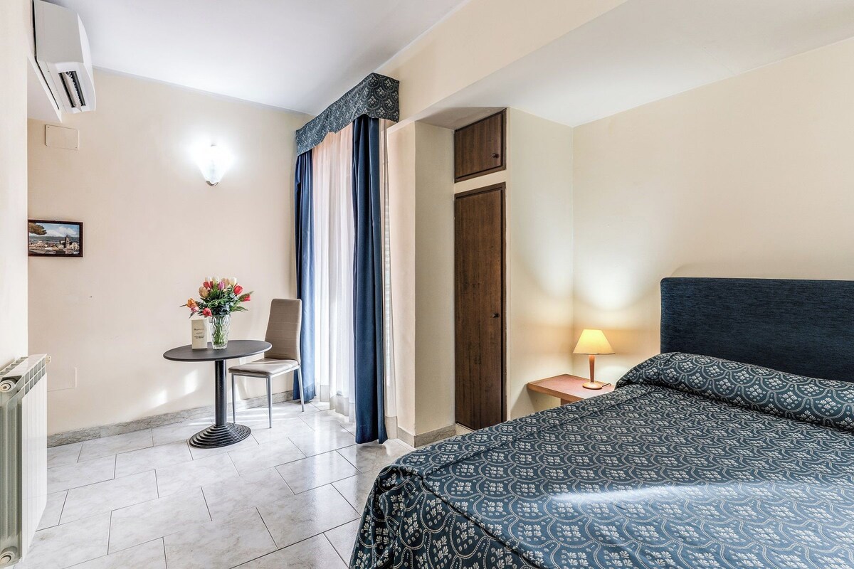 A hotel room with a bed, a small table with a lamp, a chair, and a window with curtains.