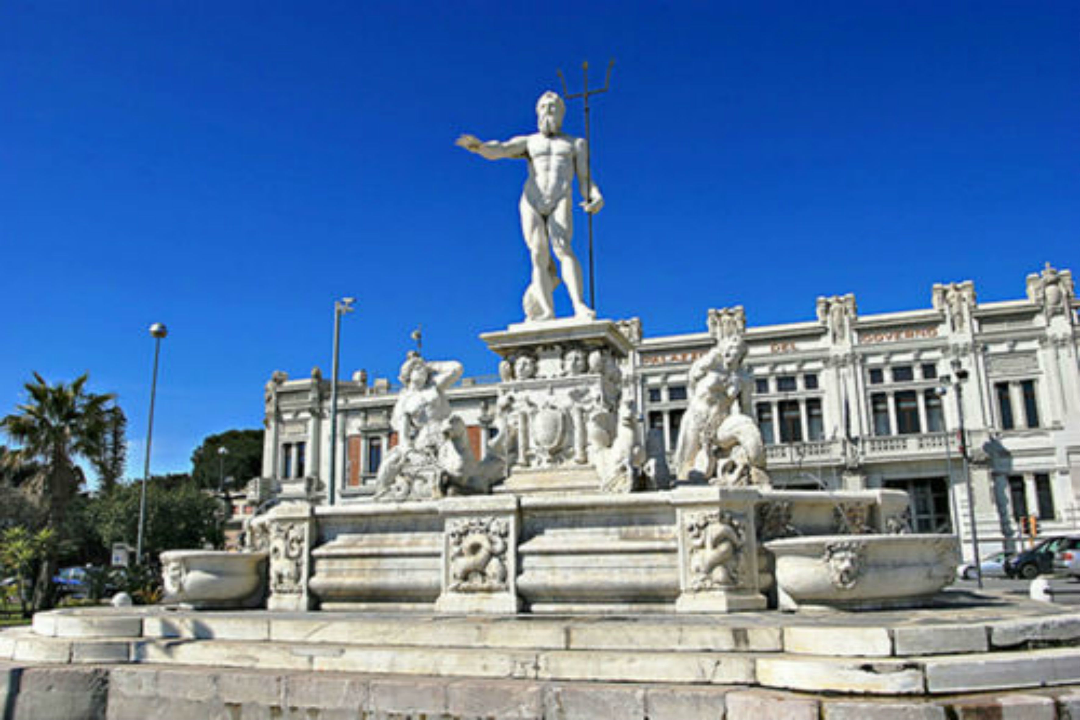 A classical statue on a fountain with ornate architectural details and a building in the background.