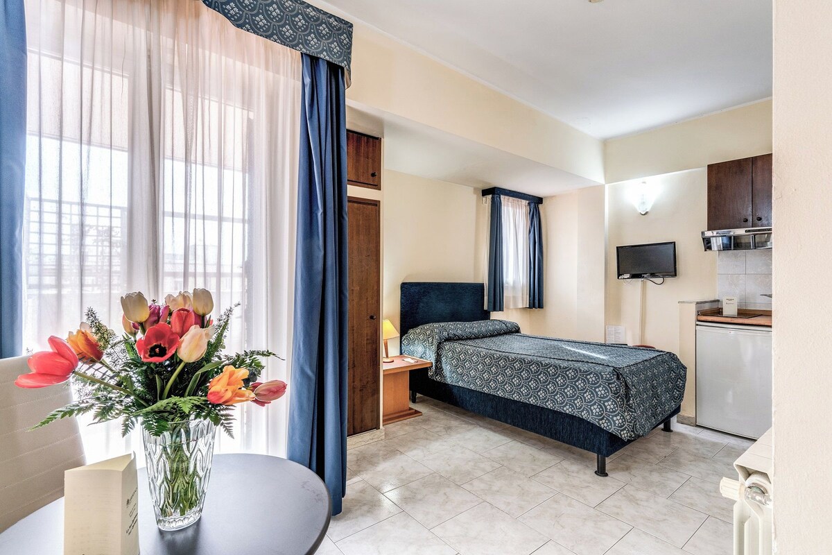 A hotel room with a bed, a small desk, a chair, a table with a vase of flowers, a TV, and a kitchenette.