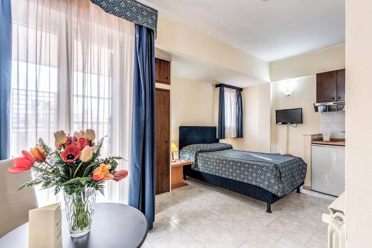 A hotel room with a bed, a small desk, a chair, a table with a vase of flowers, a TV, and a kitchenette.