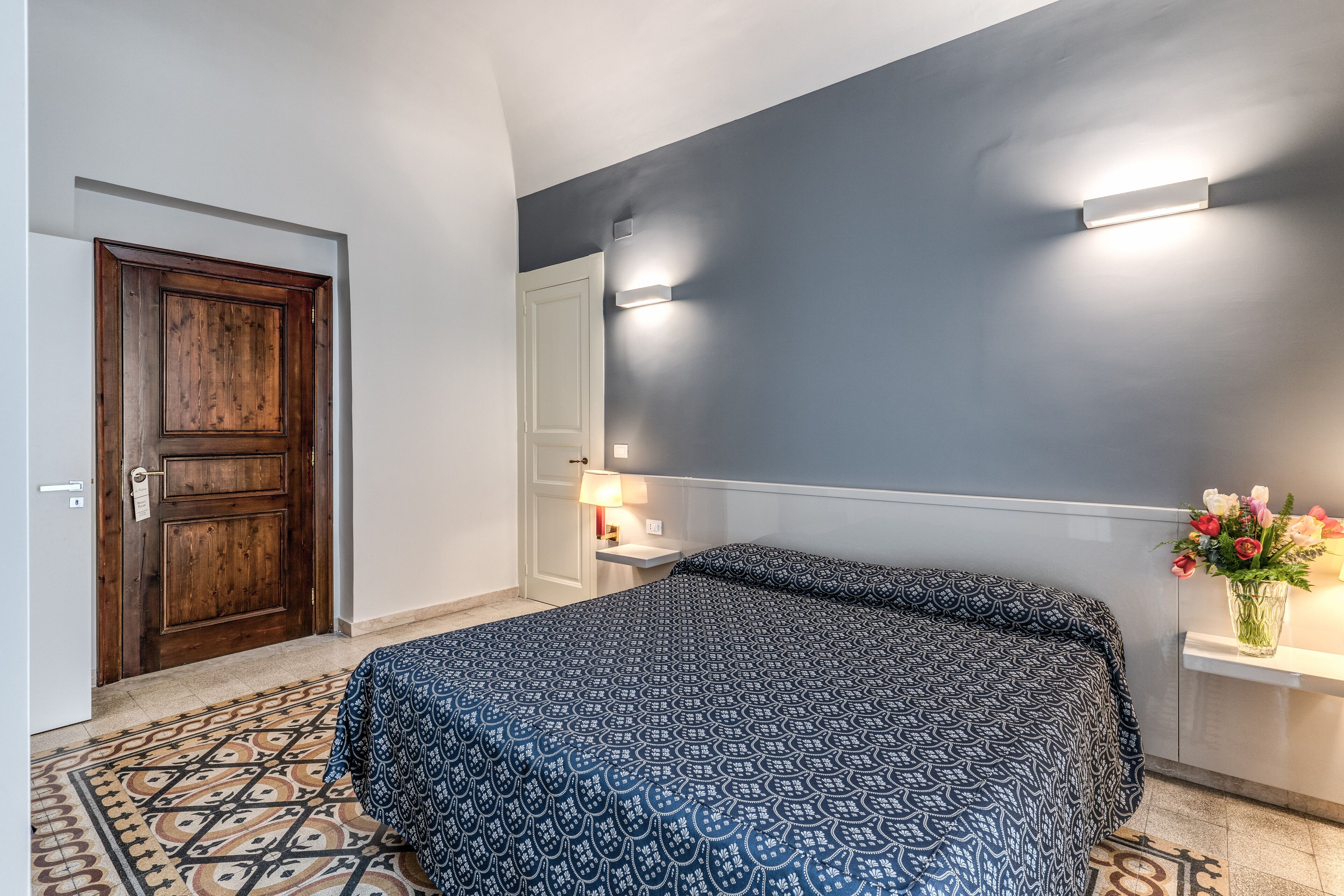 Een slaapkamer met een bed met een patroontekening, een houten deur, een nachtkastje met een lamp en een vaas met bloemen.