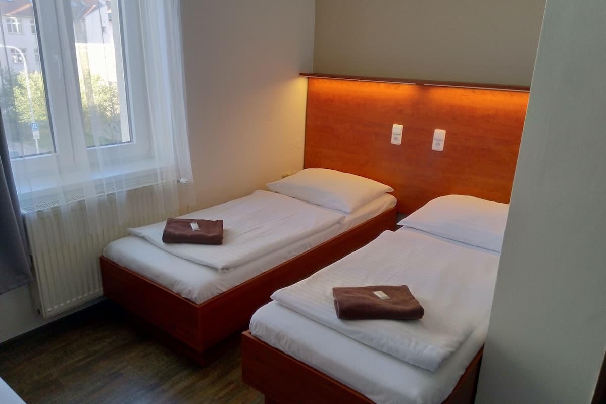 Une petite pièce bien éclairée, avec deux lits simples, chacun doté d’une tête de lit en bois et de draps blancs.