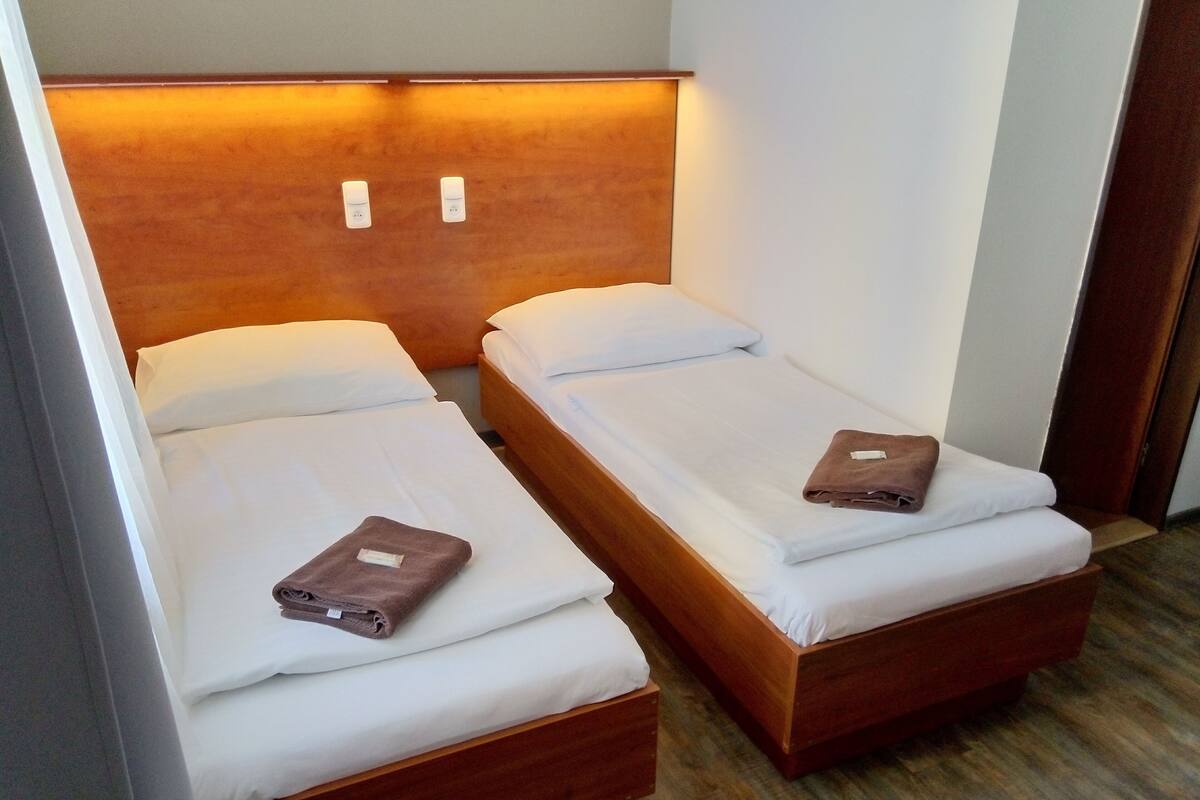 Deux lits simples avec des têtes de lit en bois et du linge de lit blanc, un plancher en bois et une petite table de chevet en bois avec une lampe.
