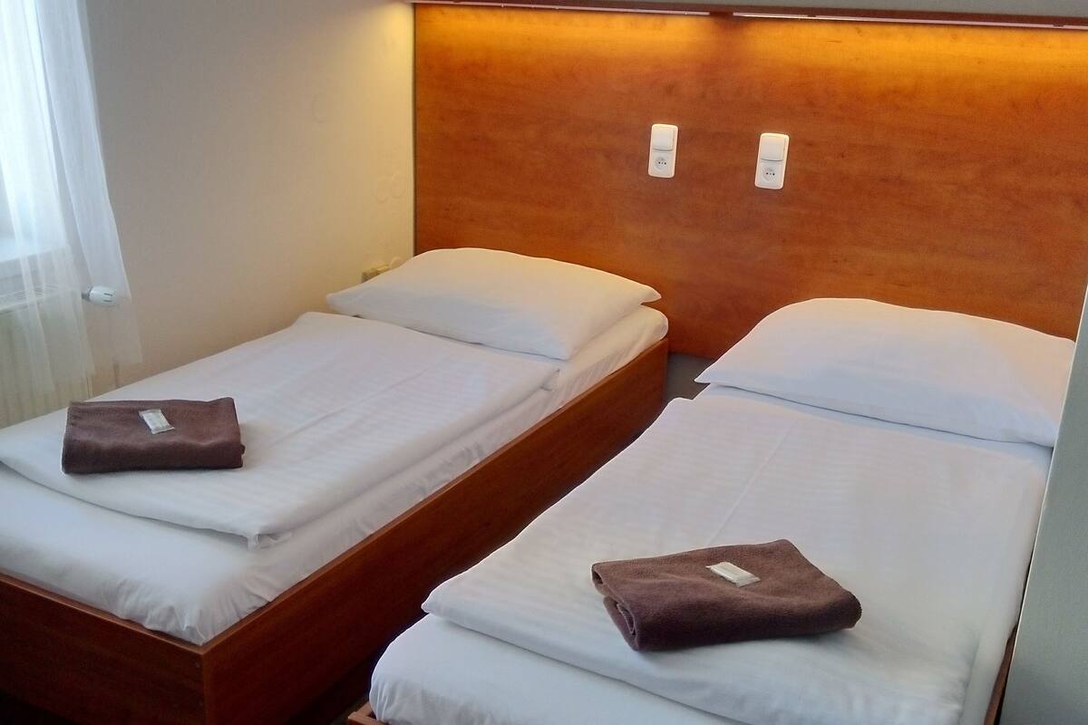 Deux lits simples avec des cadres en bois et une literie blanche, une tête de lit en bois et une prise de courant au mur.