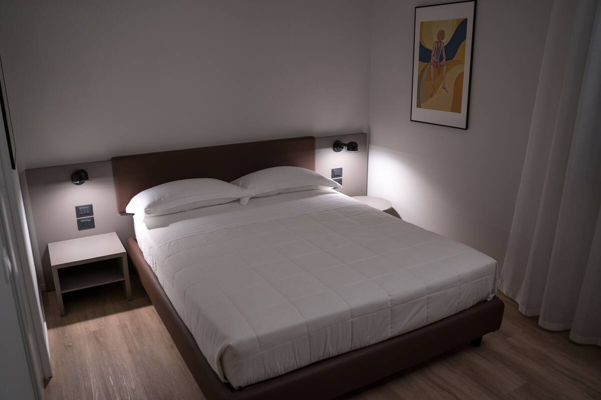 Una cama bien hecha con cabecera, mesitas de noche y una lámpara de pared.