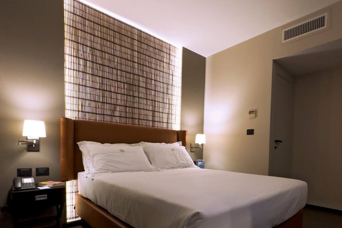 Habitación de hotel con una cama grande, dos mesitas de noche, una lámpara de pared y una ventana con persianas.