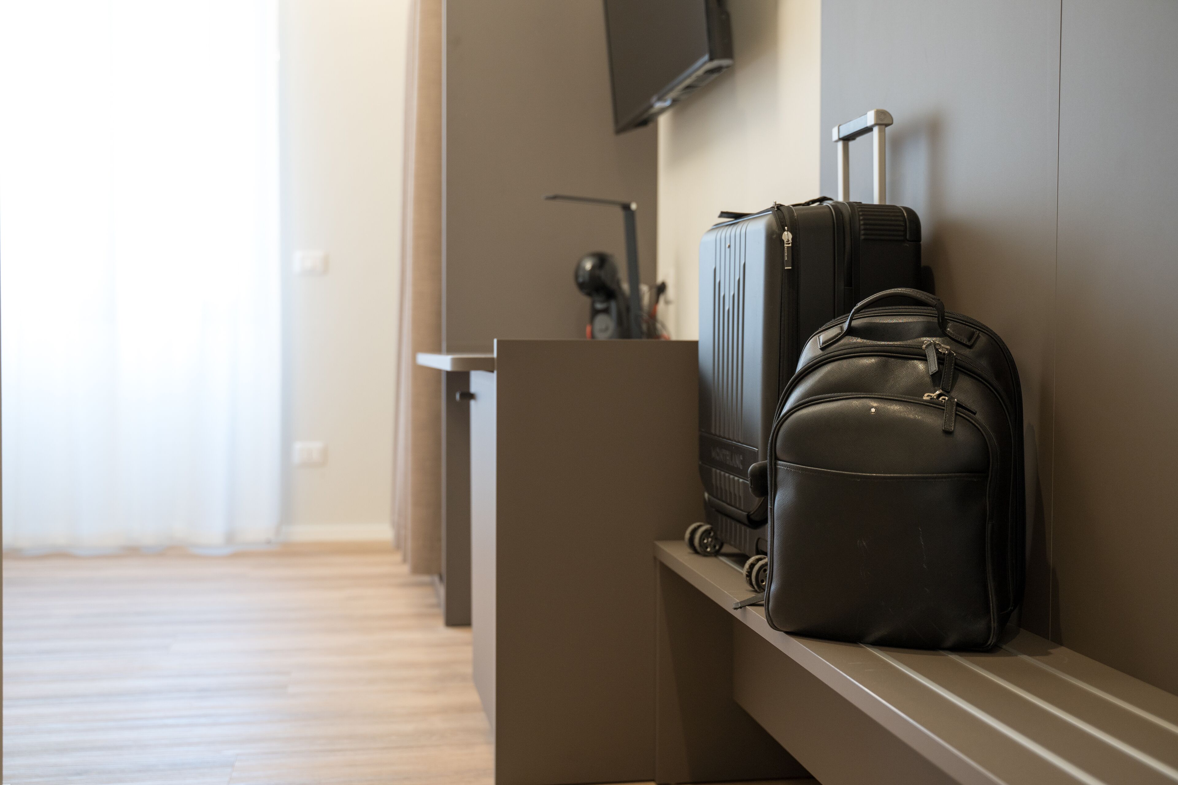 A hotel room with a wooden floor, a TV mounted on the wall, and luggage on a shelf.