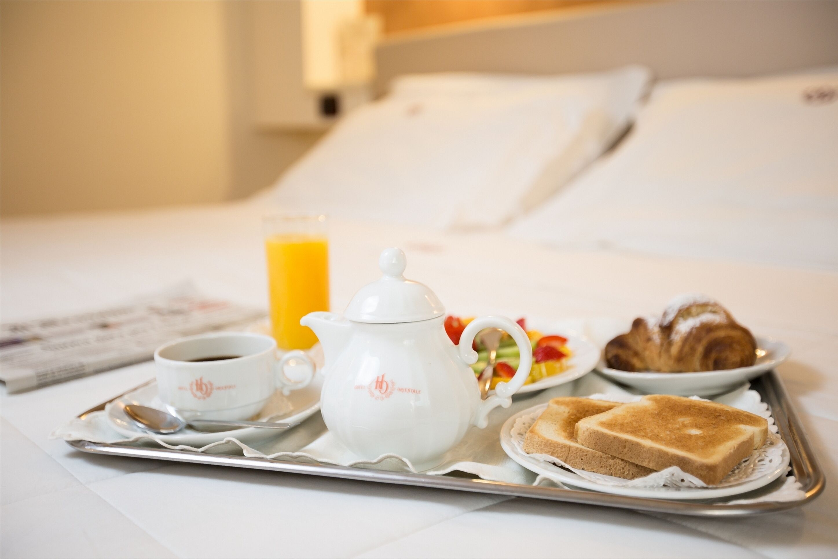 A breakfast tray with a teapot, cup, orange juice, croissant, and toast on a bed.