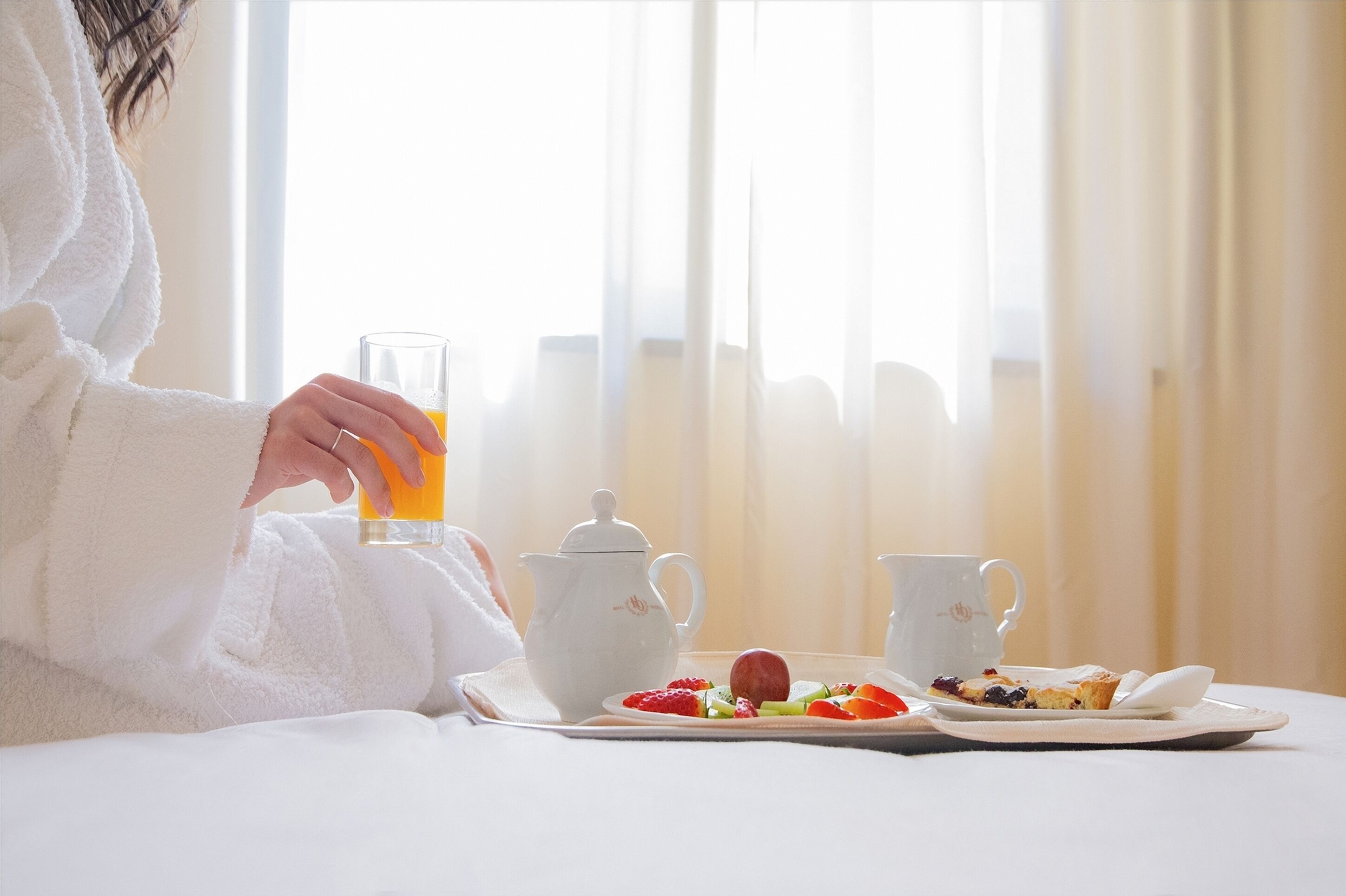 A person in a white robe sitting at a table with a glass of orange juice, a teapot, a cup, and a plate with fruit and a pastry.