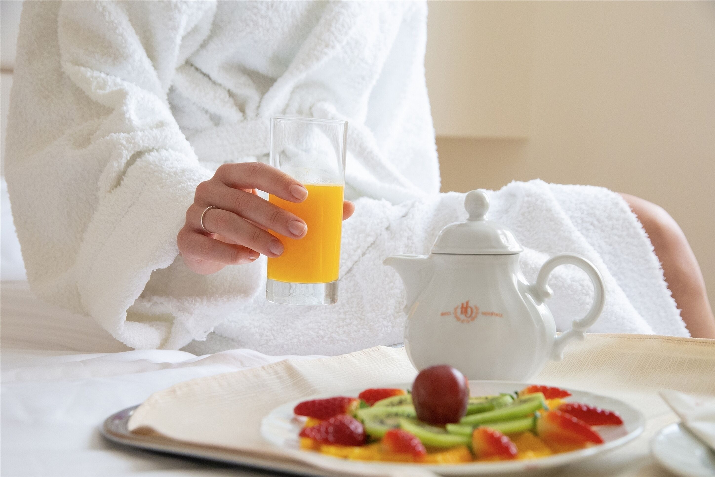 A person in a white robe holding a glass of orange juice, a white teapot, and a plate of fruit on a table.