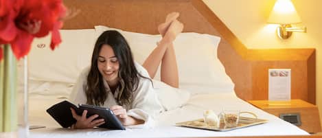 A person lying on a bed with a tablet, a tray with a glass and a small bowl, and a nightstand with a lamp and a menu.