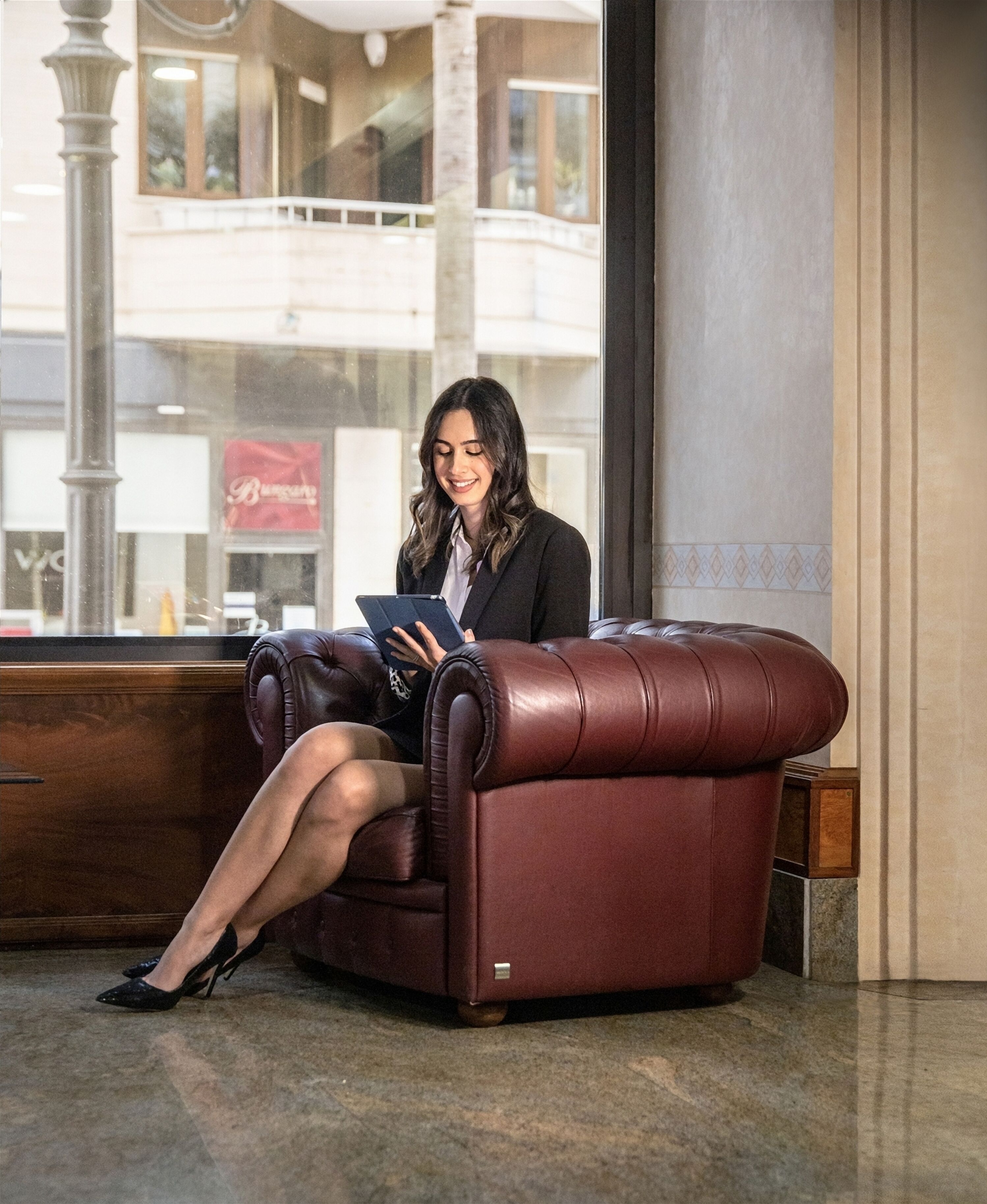 Eine Frau sitzt auf einer Ledersofa in einer Lobby mit einem großen Fenster, einer Holzbank und einem Marmorboden.