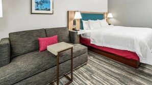 Une chambre d’hôtel avec un canapé gris, une petite table, un lit avec une literie blanche et un tableau encadré au mur.