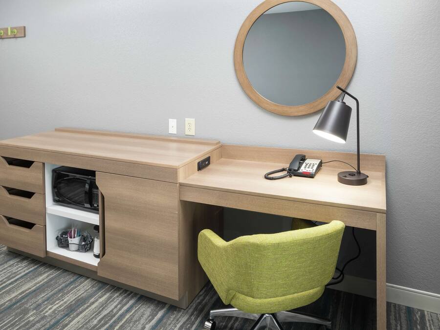Een moderne hotelkamer met een houten bureau, een groene stoel, een ronde spiegel en een bureaulamp.
