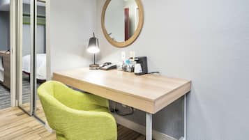 A hotel room with a desk, a mirror, a green chair, a bed, and a wooden floor.