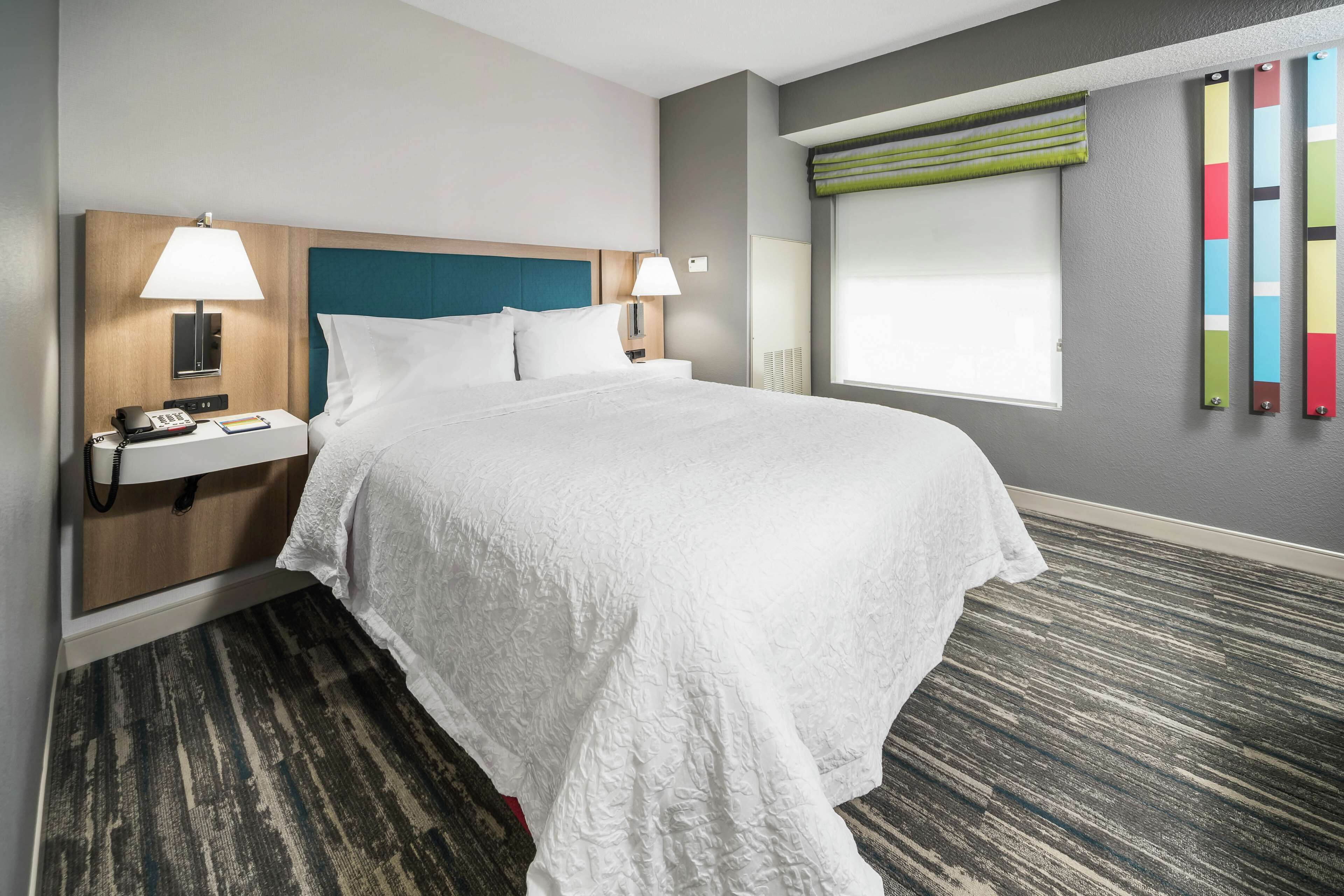A hotel room with a large bed, two bedside tables with lamps, a headboard, a window with blinds, and a carpeted floor.