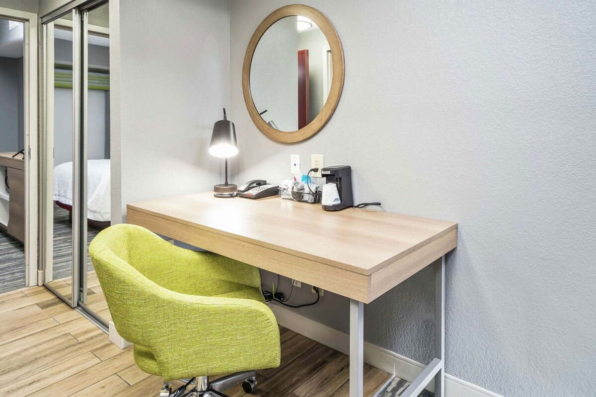 Una habitación de hotel con escritorio, espejo, lámpara, silla y cama.