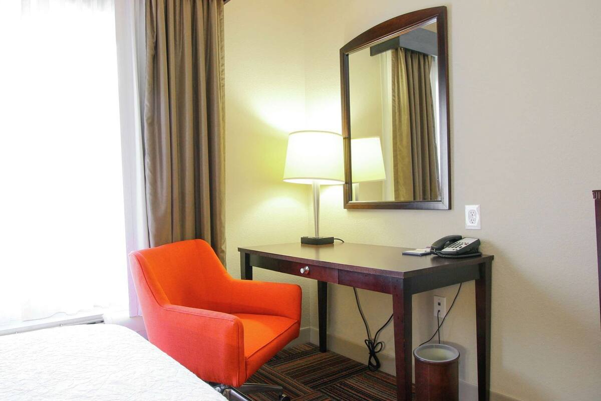 A hotel room with a bed, an orange chair, a desk with a lamp, a phone, and a mirror.