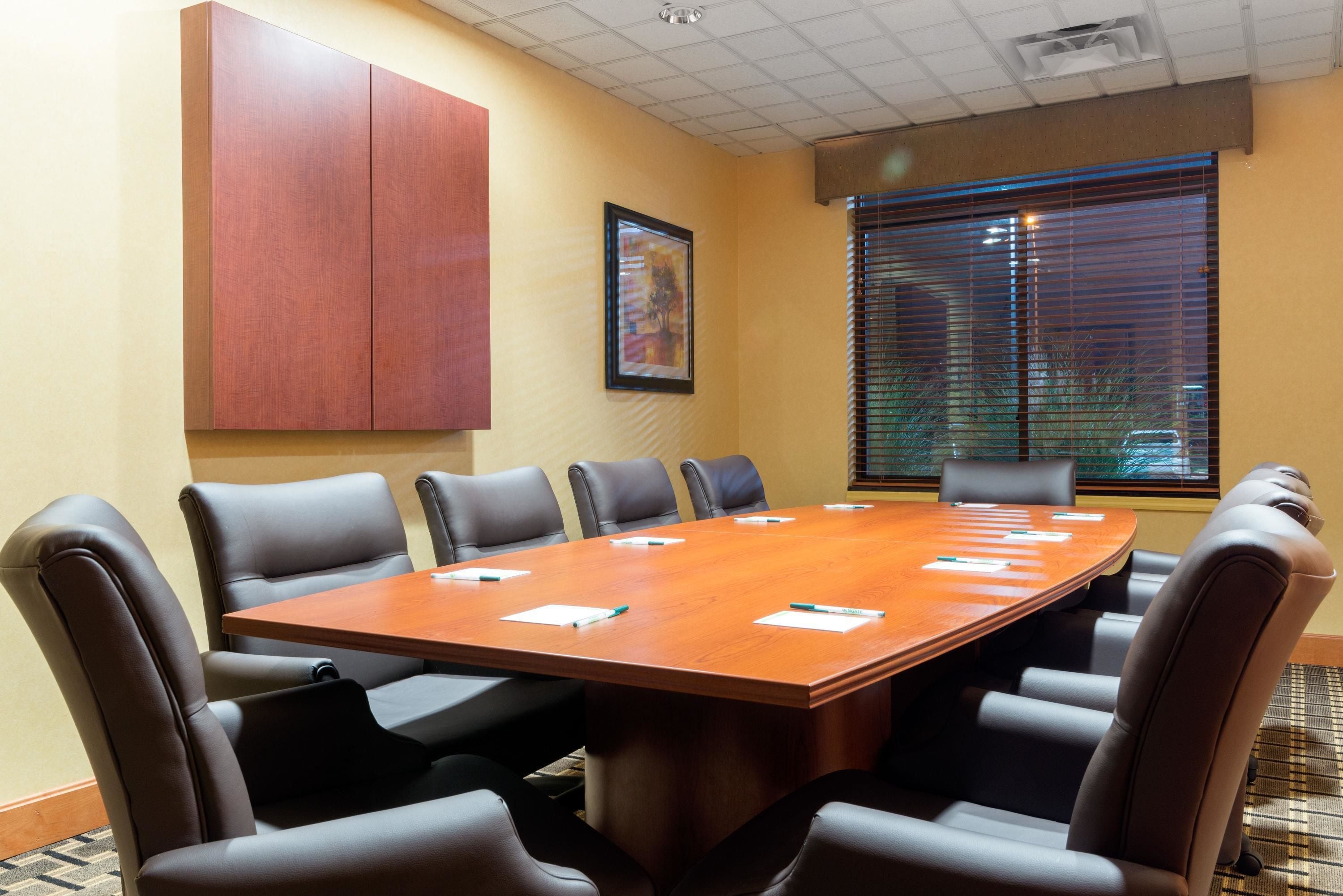 A conference room with a large wooden table, eight chairs, a red cabinet, a framed picture, and a window with blinds.