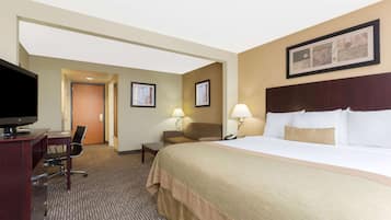 Habitación de hotel con una cama grande, un escritorio con televisor, dos sillones, una lámpara y un cuadro en la pared.