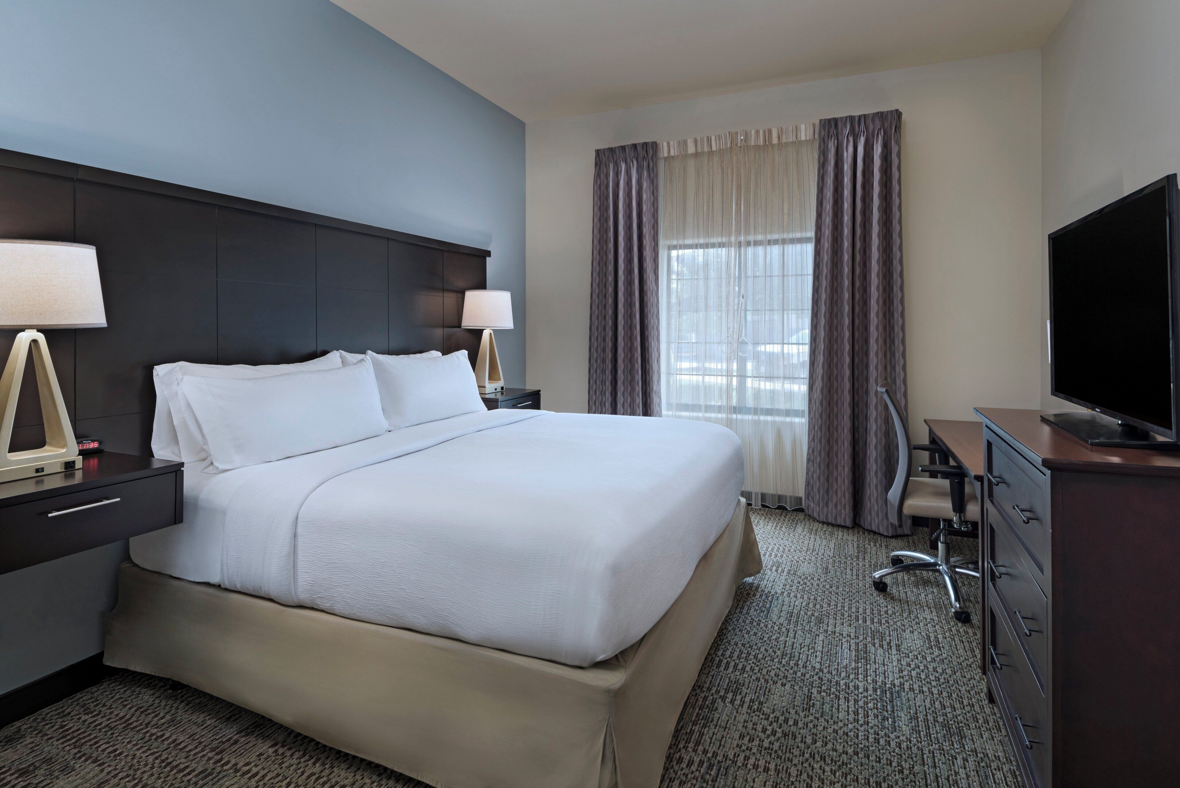 A hotel room with a large bed, two bedside tables with lamps, a TV, a desk with a chair, and a window with curtains.