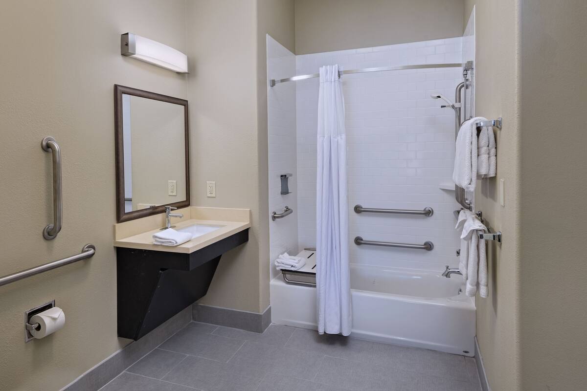 A bathroom with a sink, mirror, towel rack, shower, and grab bars.