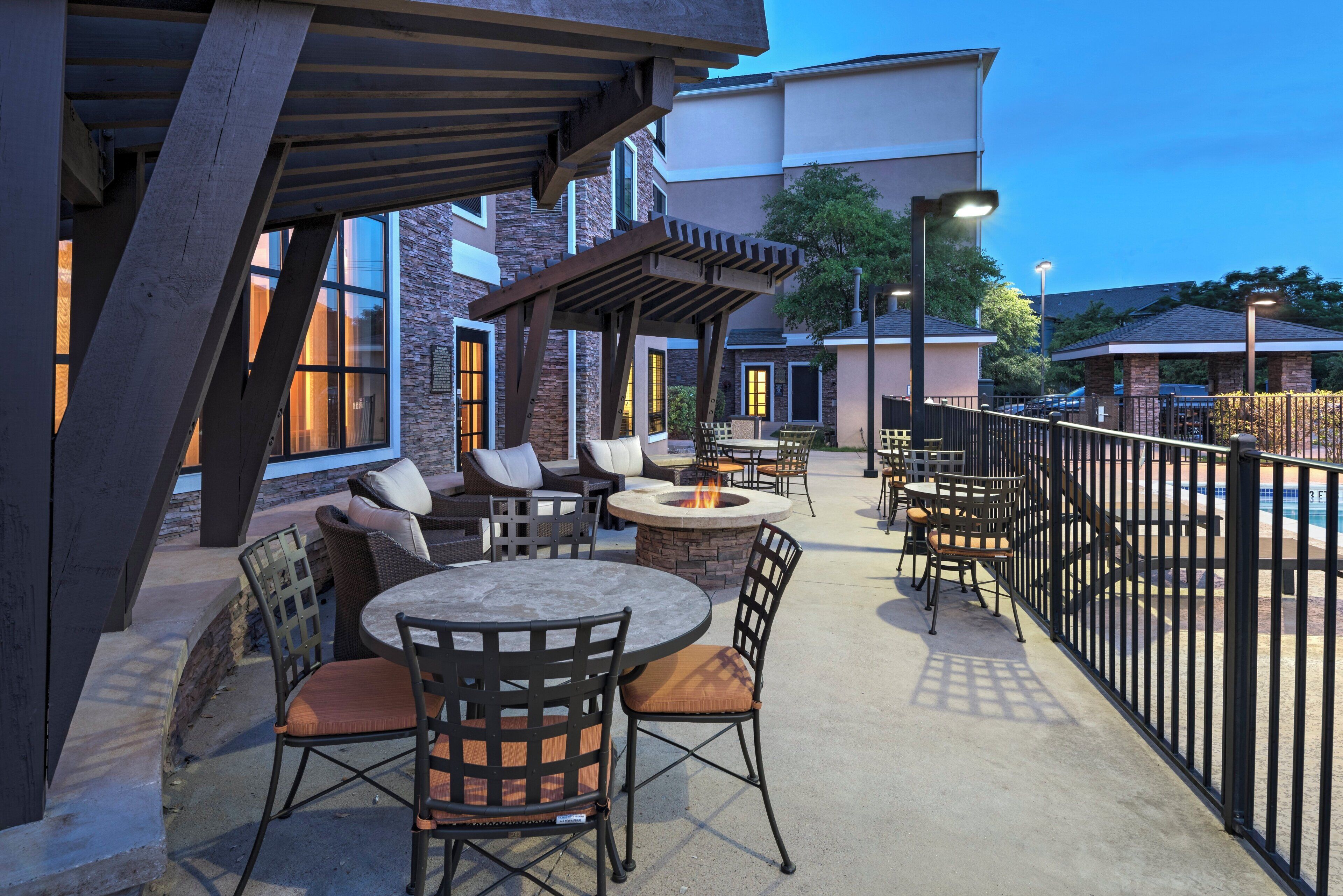 An outdoor patio area with round tables and chairs, a fire pit, and a view of a building in the background.