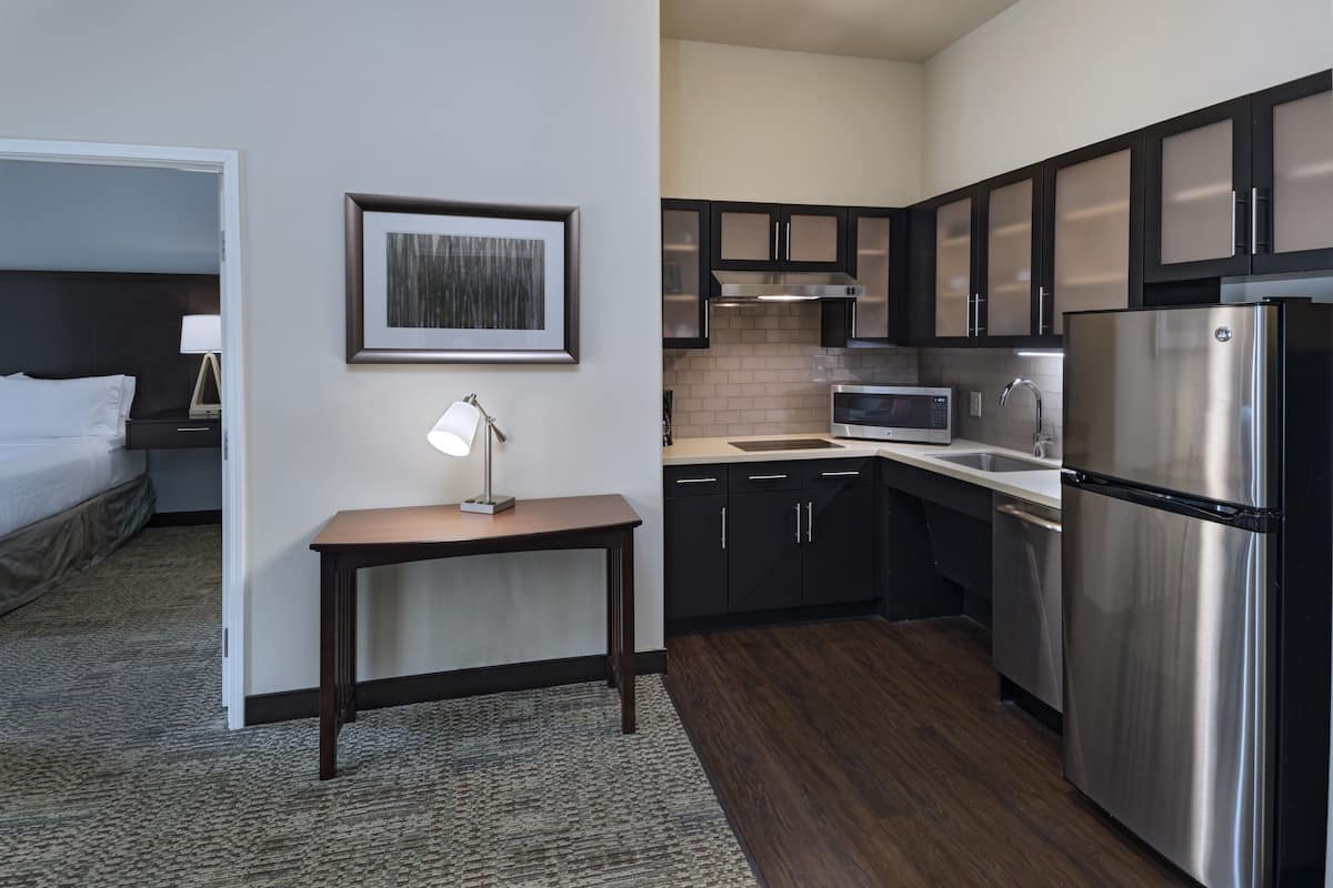 A modern hotel room with a kitchenette, a bed, a bedside table with a lamp, and a framed picture on the wall.
