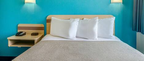 Une chambre d’hôtel avec un mur de couleur turquoise, une tête de lit en bois, un lit avec une literie blanche, deux lampes de chevet, une table de chevet avec un téléphone et une horloge, et une fenêtre avec des rideaux.