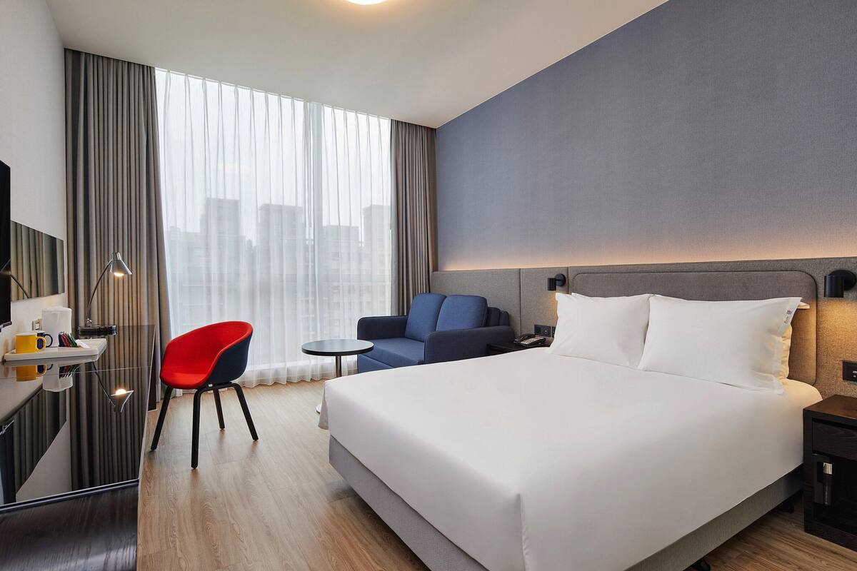 Camera d'albergo con un letto grande, una scrivania con televisore, una poltrona rossa, un tavolino e vista sulla città dalla finestra.