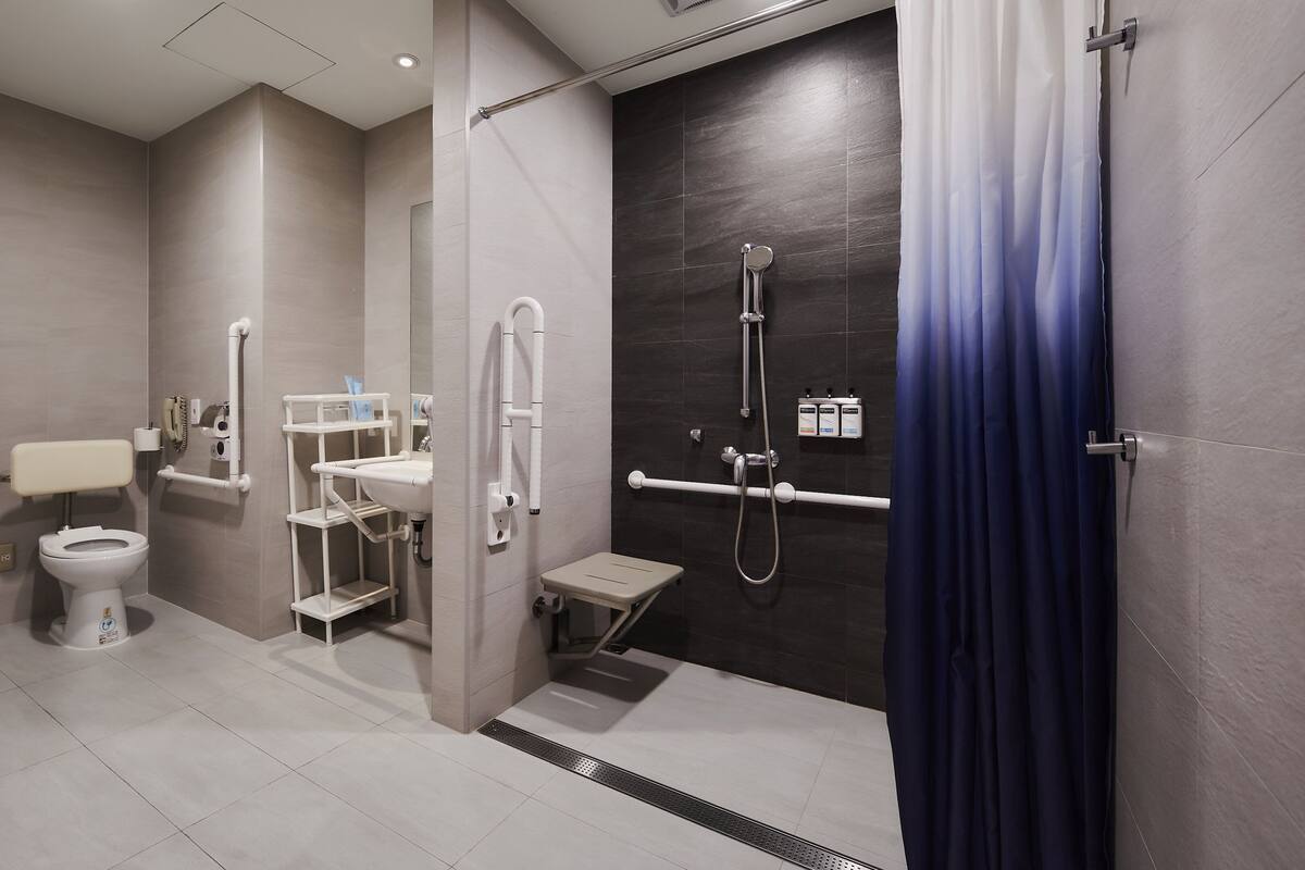 Un bagno moderno dotato di toilette, lavabo e zona doccia, con maniglie di sostegno e un poggiapiedi.