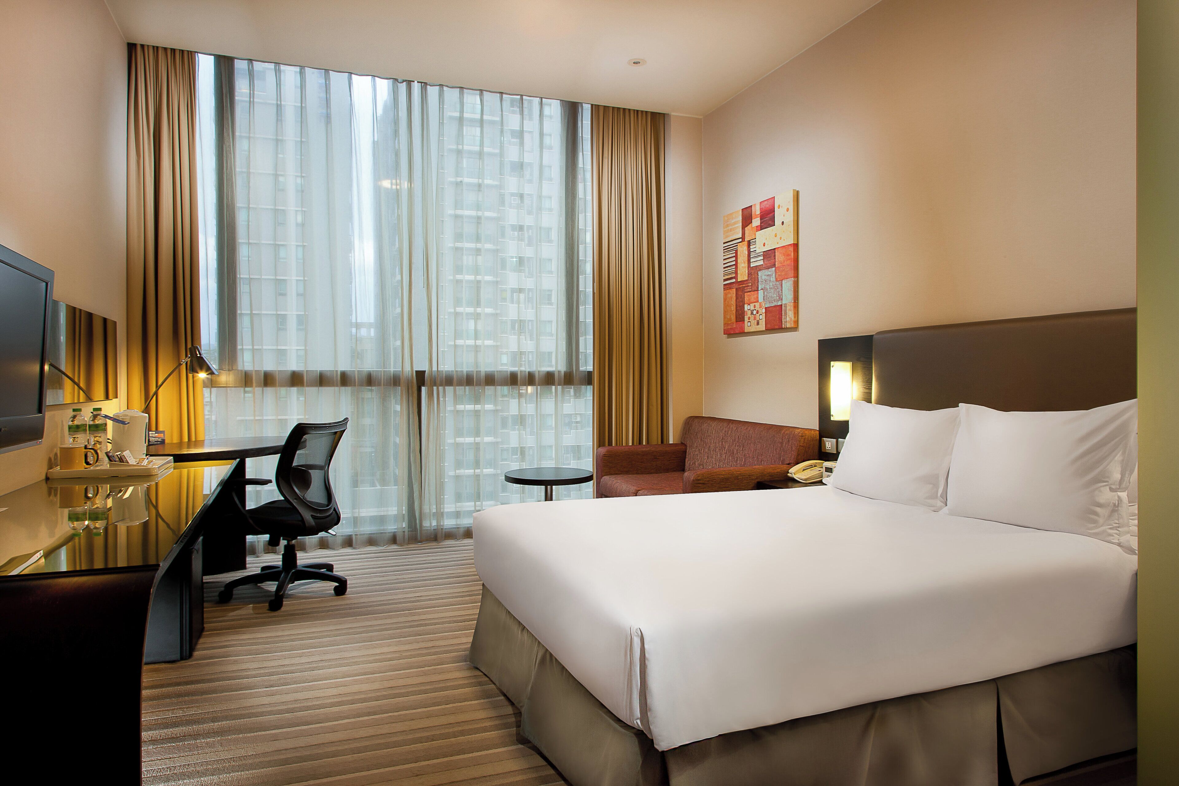 A hotel room with a bed, a desk with a chair, a small table, a telephone, and a window with curtains.