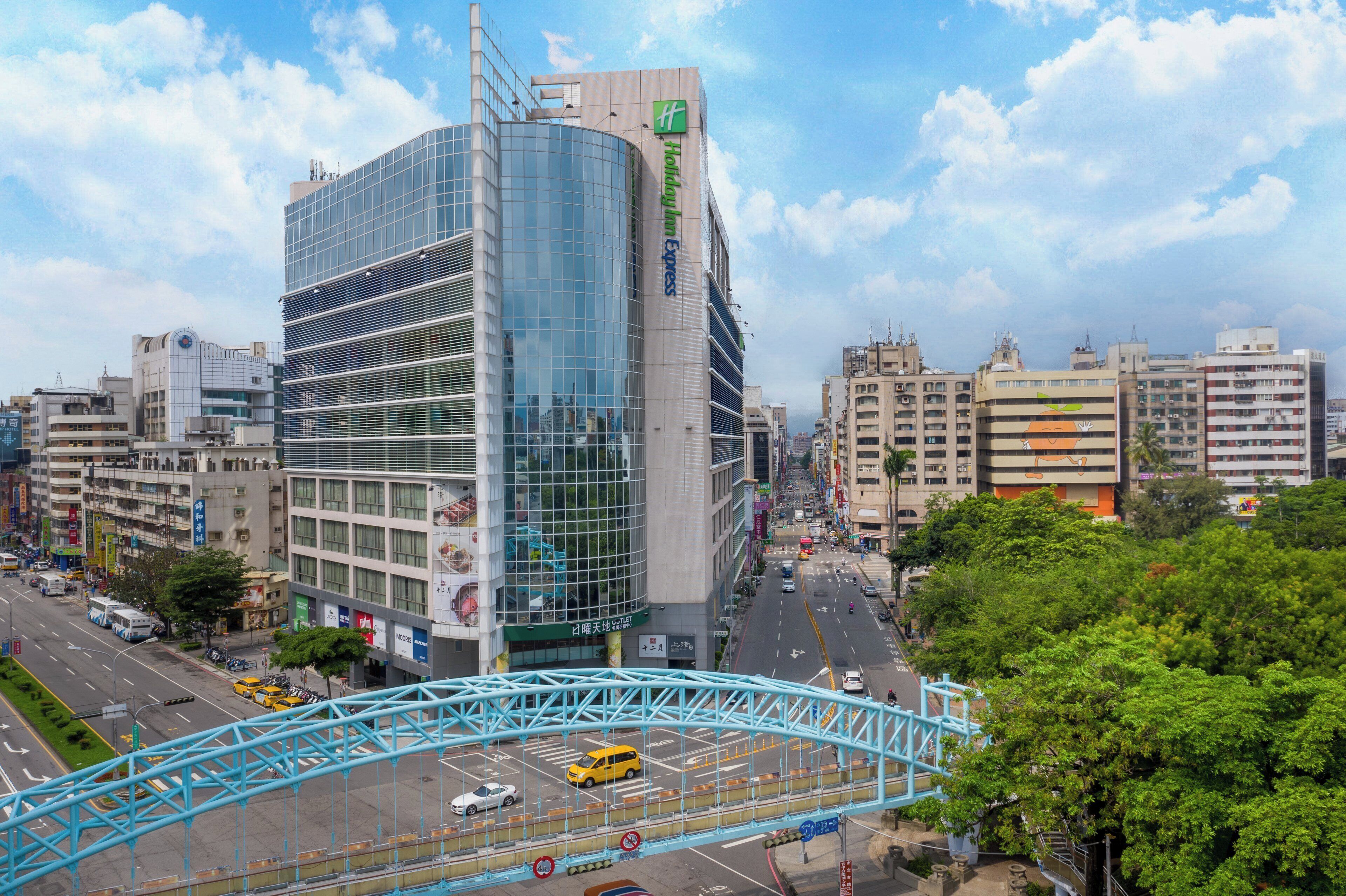 Un edificio moderno con fachada de vidrio, rodeado por una concurrida calle urbana con vehículos y un puente peatonal.