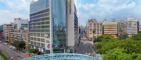 Un edificio moderno con fachada de vidrio, rodeado por una concurrida calle urbana con vehículos y un puente peatonal.