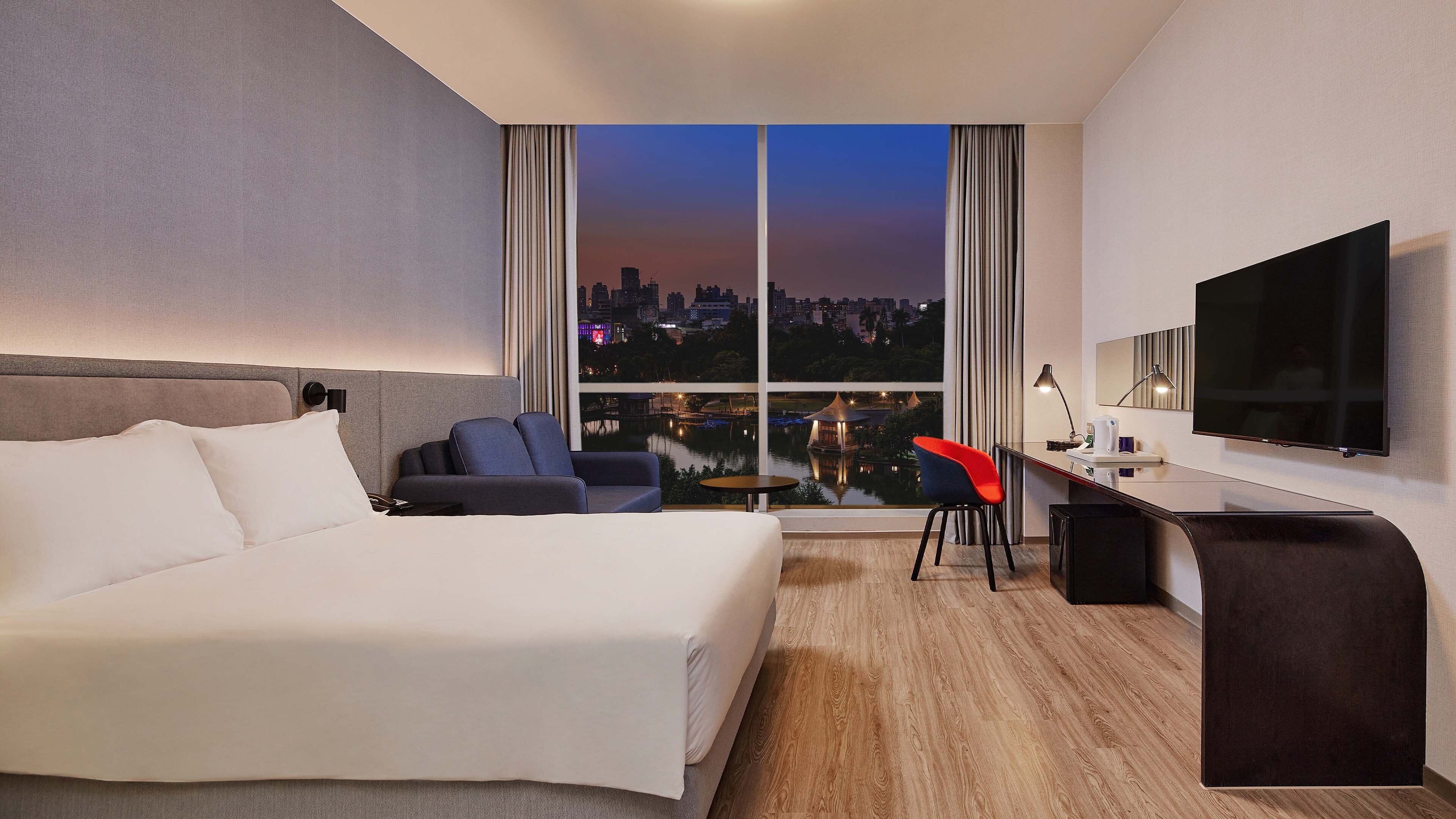 A hotel room with a bed, a desk, a chair, a TV, and a view of a cityscape at dusk.