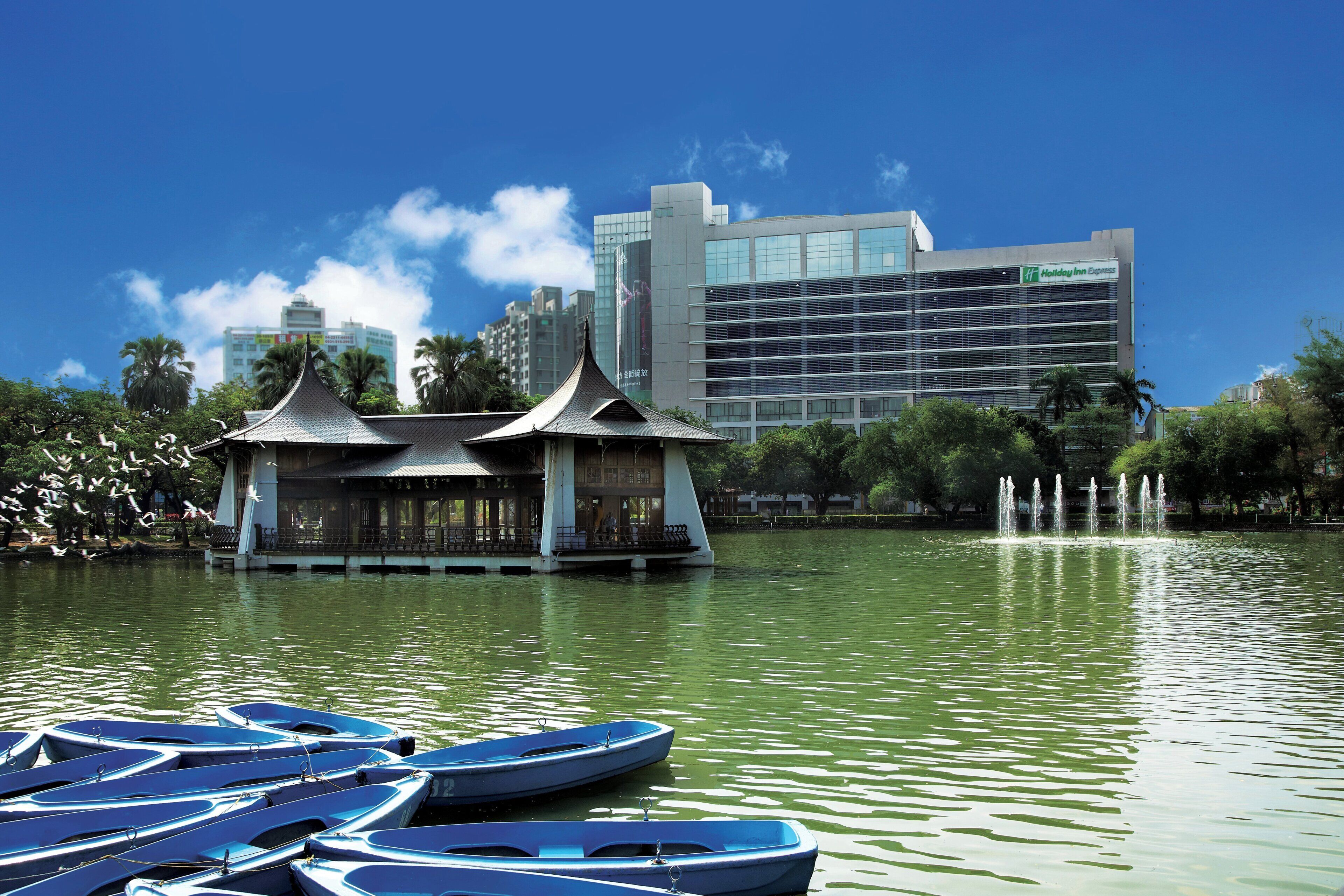 Ein traditioneller Pavillon am See mit Booten, im Hintergrund ein Holiday Inn Express Gebäude.