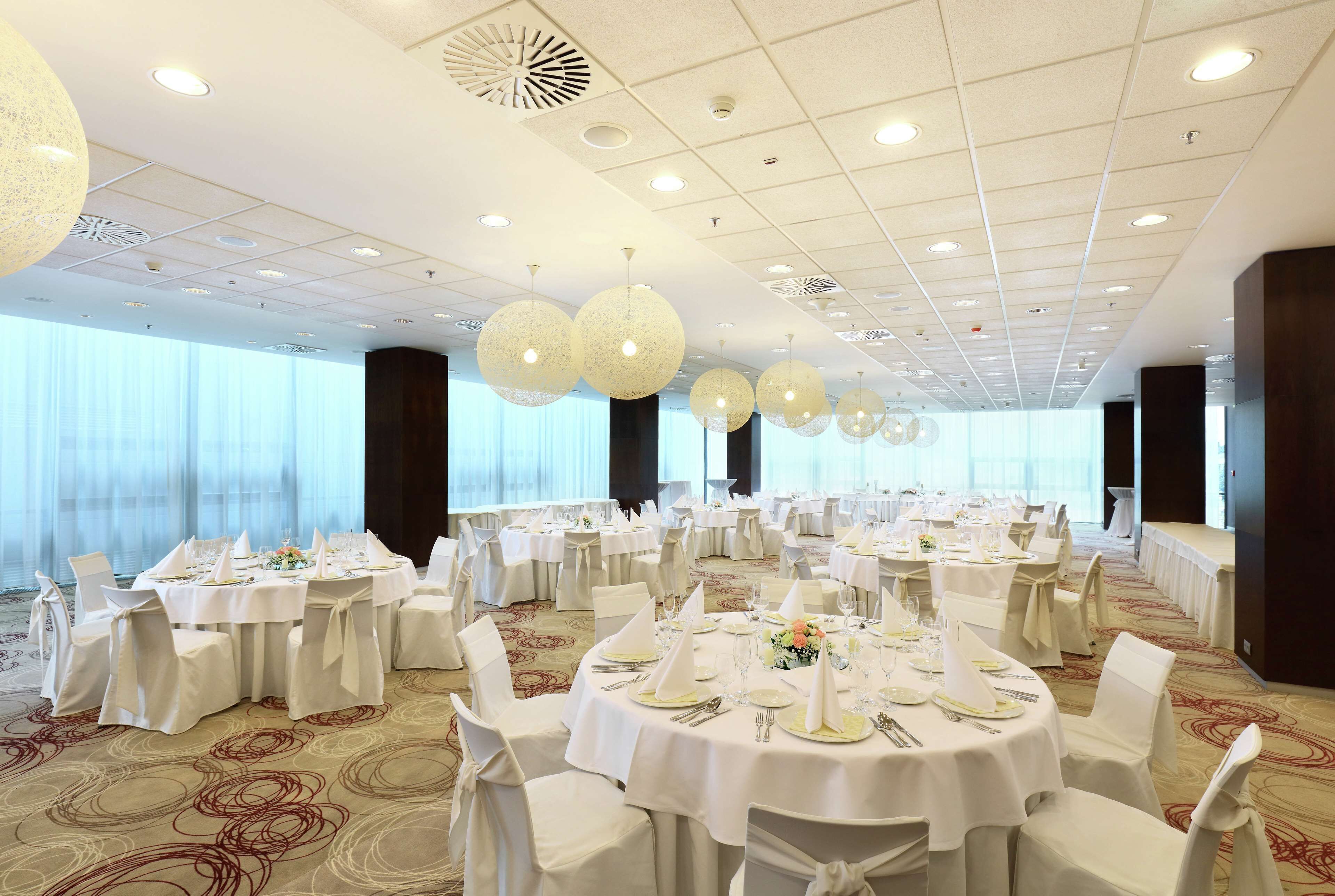 Una sala de banquetes amb taules rodones preparades per dinar, grans finestres i un sostre alt amb il·luminació empotrada.