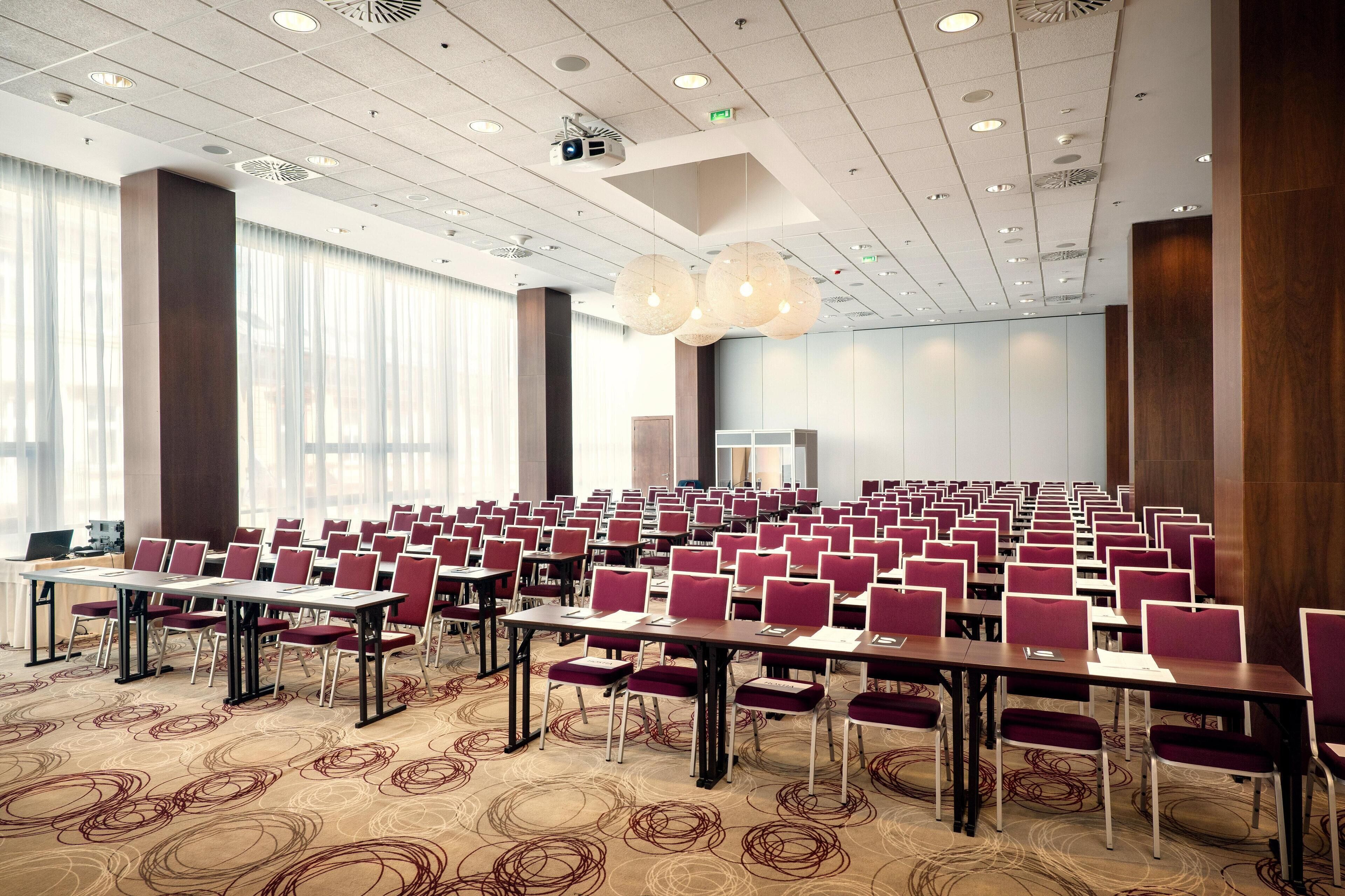 A conference room with rows of tables and chairs, large windows, and a ceiling with recessed lighting and a projector.
