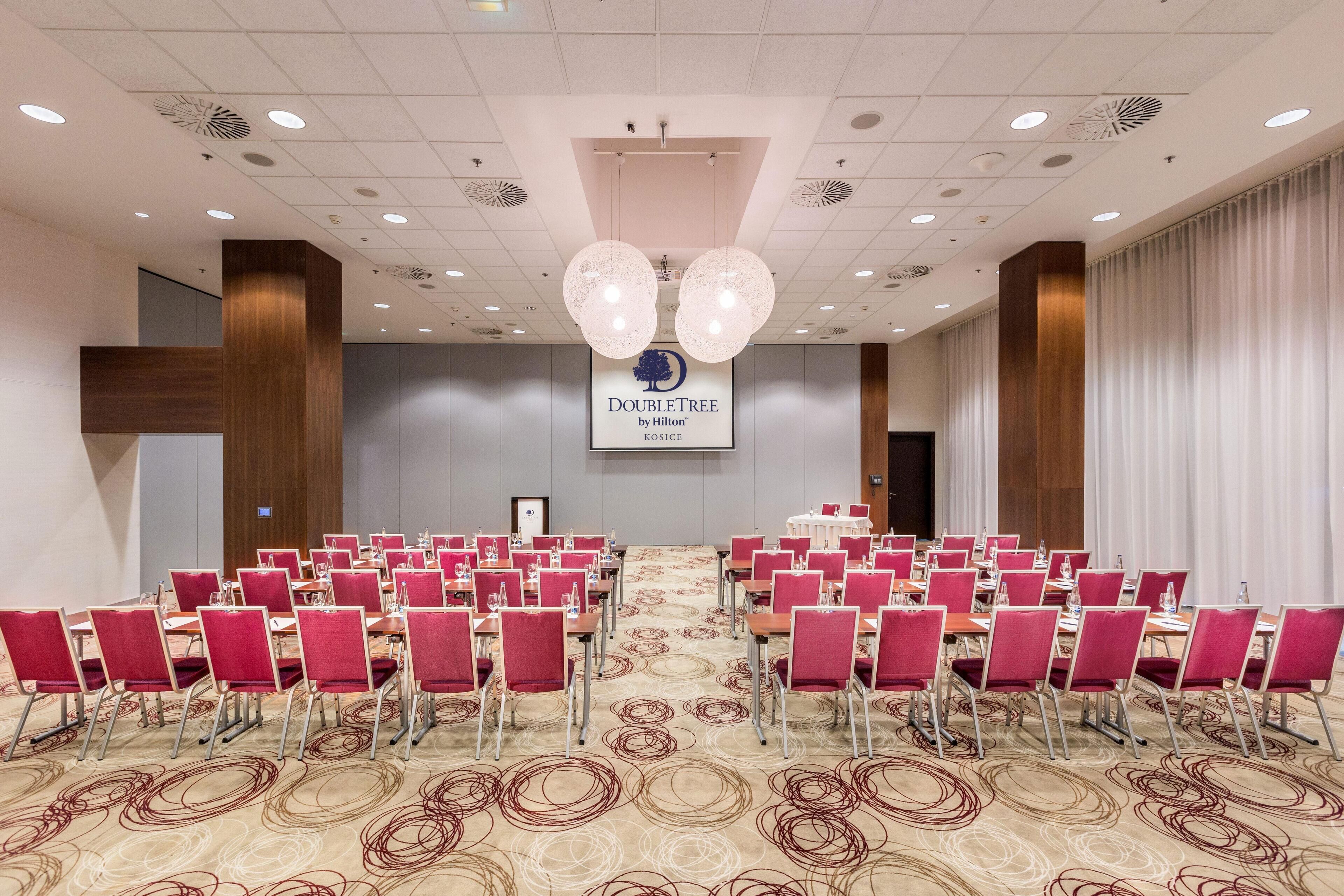 A conference room with rows of red chairs, a patterned carpet, and a ceiling with recessed lighting and a central chandelier.