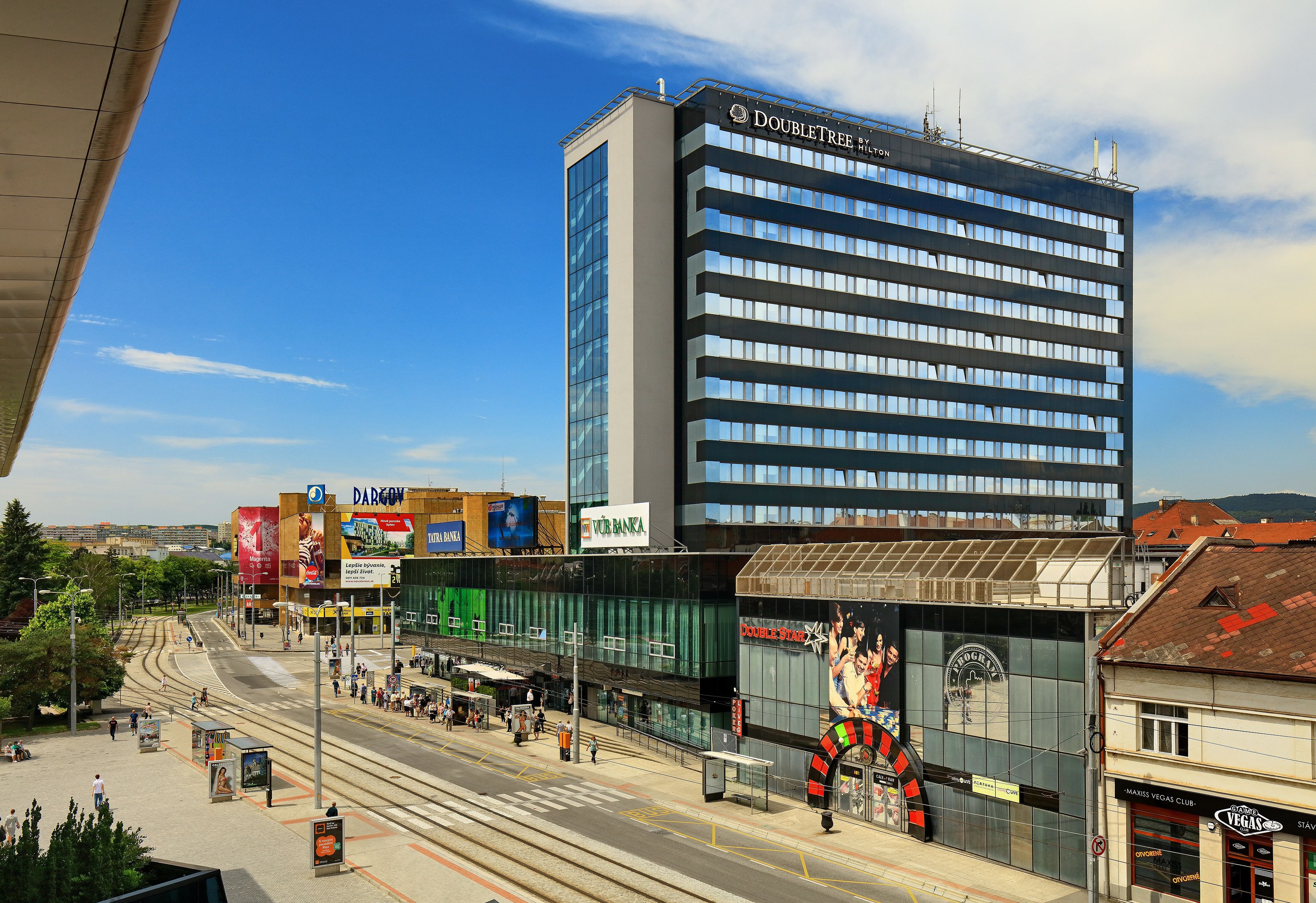 Un hôtel moderne, reconnaissable à son enseigne bien visible, situé près d’une ligne de tramway et d’une rue animée bordée de divers commerces.