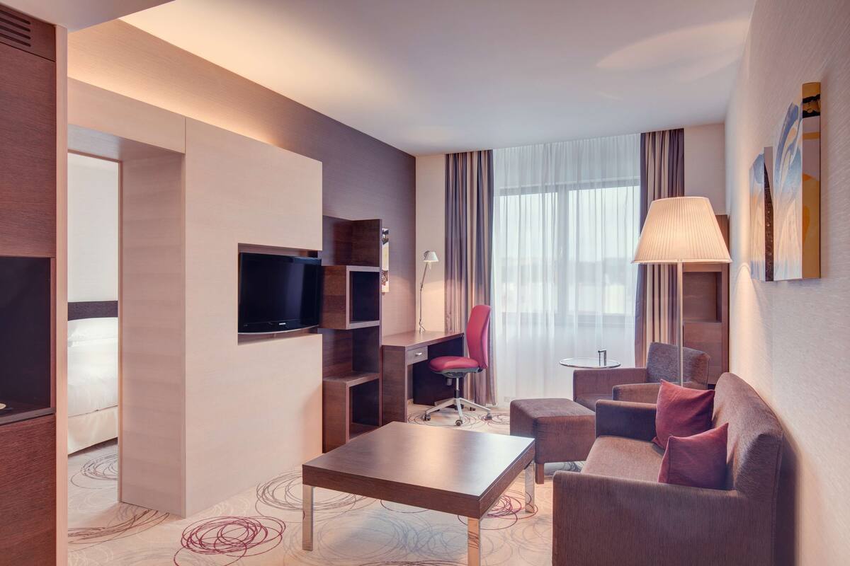 A modern hotel room with a flat-screen TV, a desk, a red office chair, a sofa, and a lamp.
