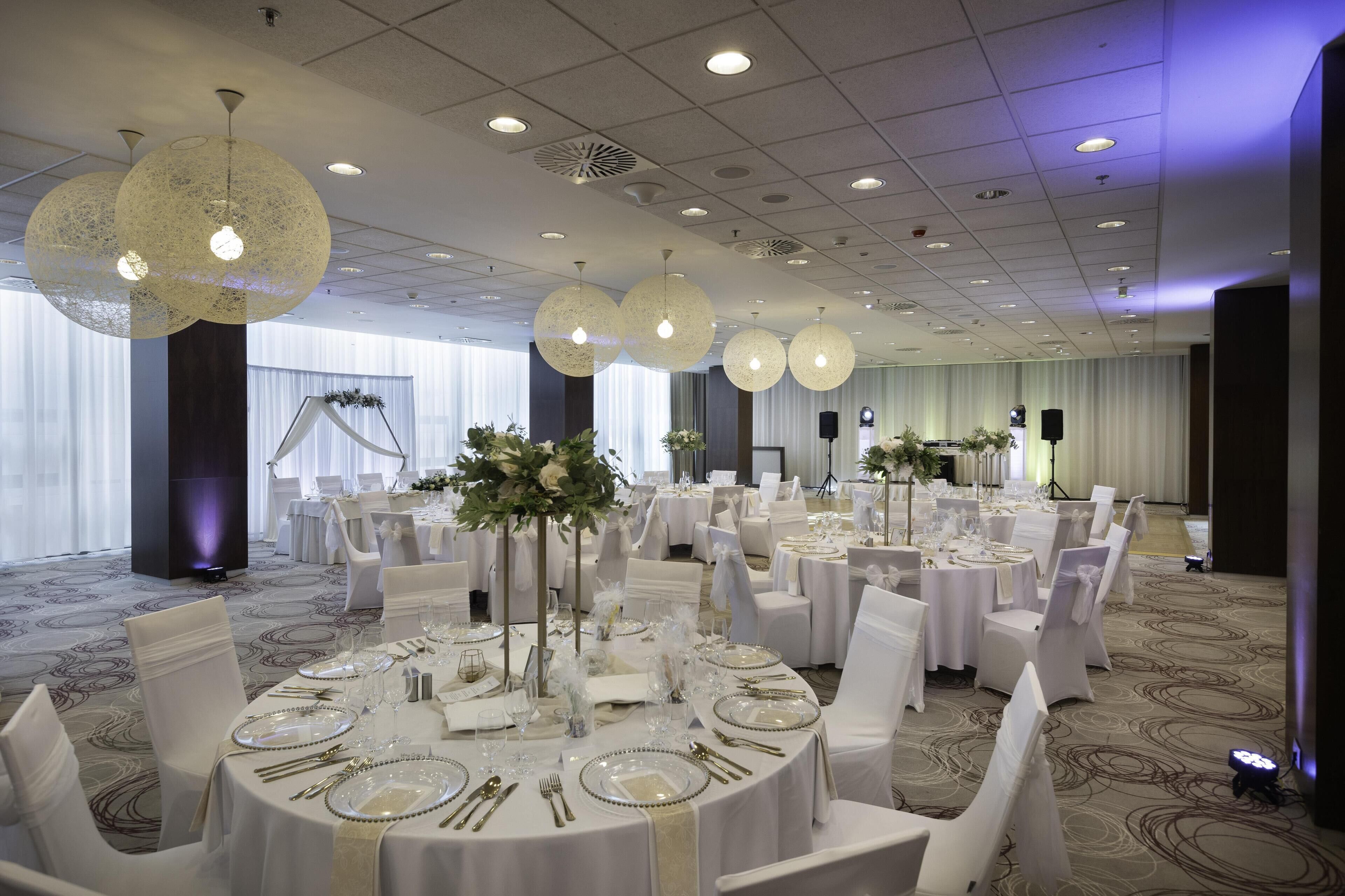 Un salón de banquetes con mesas redondas dispuestas para cenar, sillas blancas y centros de mesa decorativos.